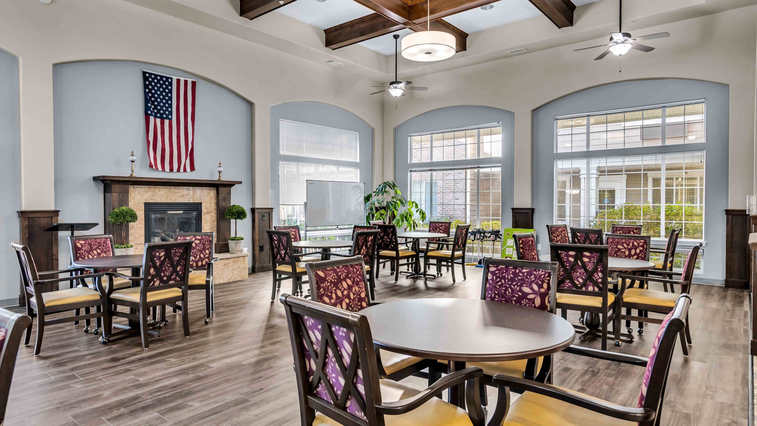 Spacious, well-lit dining room with round tables and patterned chairs, large windows, a fireplace with an American flag above it, and greenery throughout.