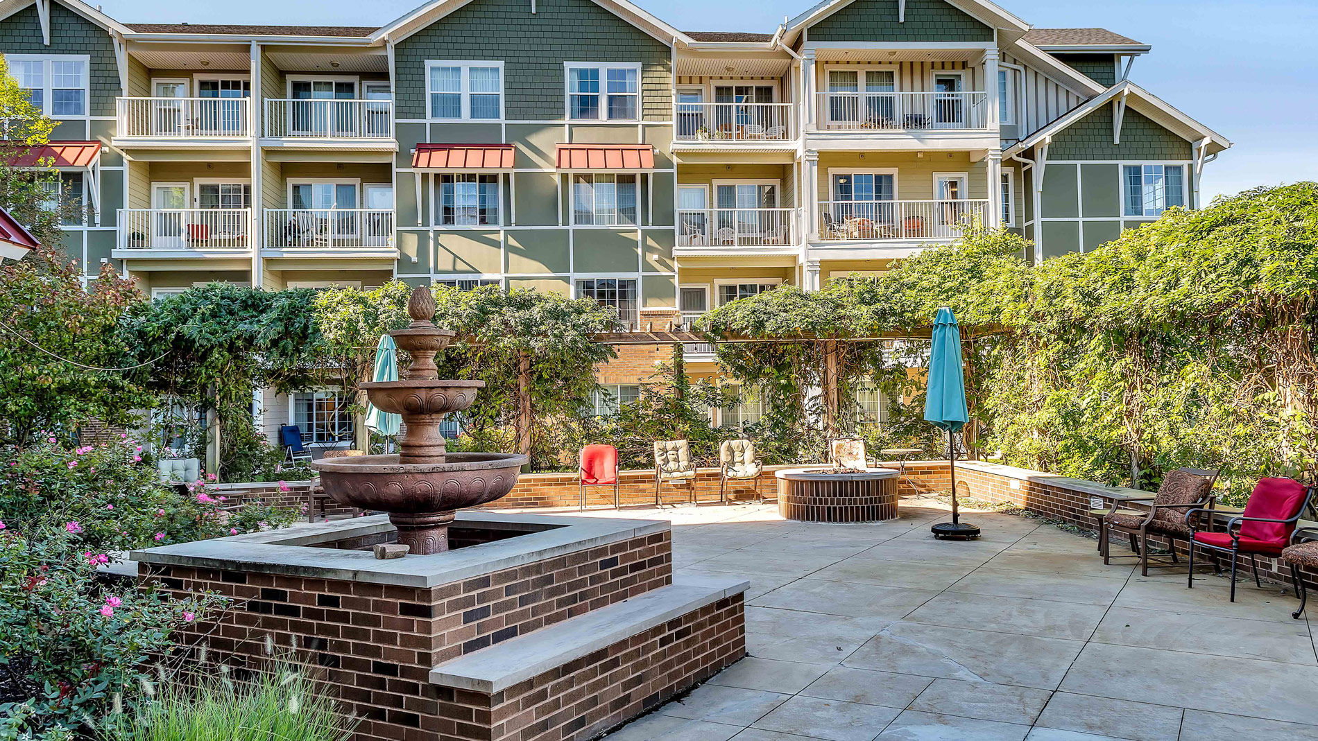 A courtyard with a brick fountain, patio seating, a round fire pit, and green trellis-covered walls, set in front of a multi-story residential building with balconies.