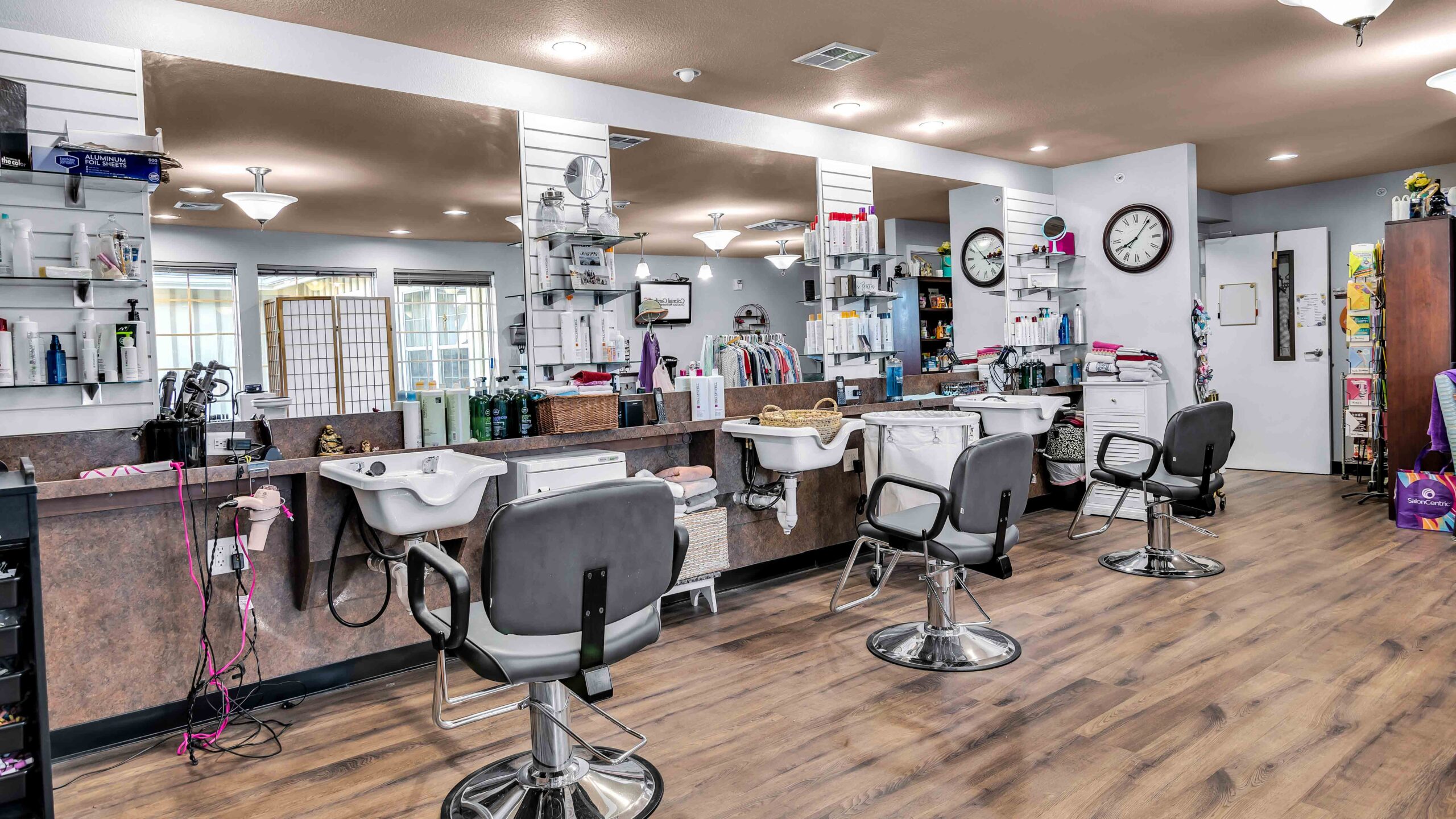 A modern hair salon with three styling stations, mirrors, swivel chairs, and shelves holding various hair products and tools, all set on wood flooring under bright lighting.
