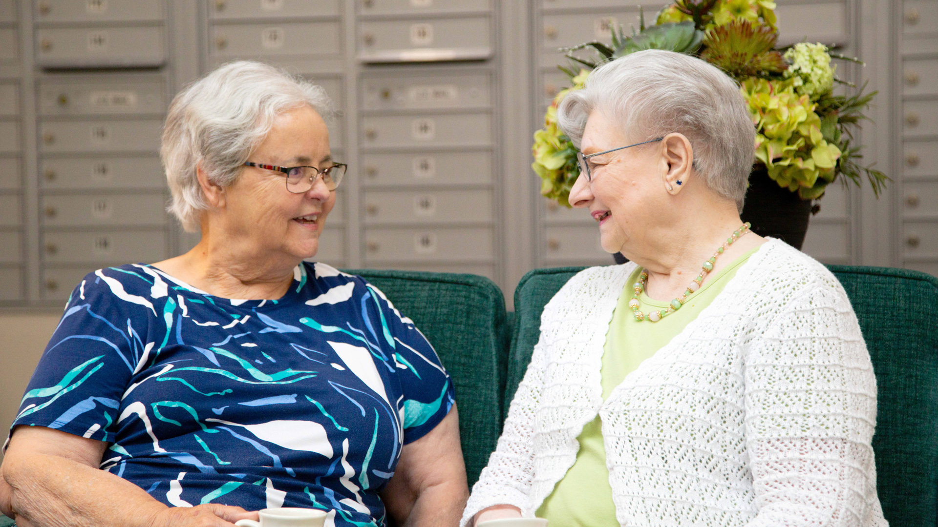 Two elderly women sit on a green sofa, smiling and talking to each other. They appear relaxed and friendly, with mailboxes and a flower arrangement visible in the background.