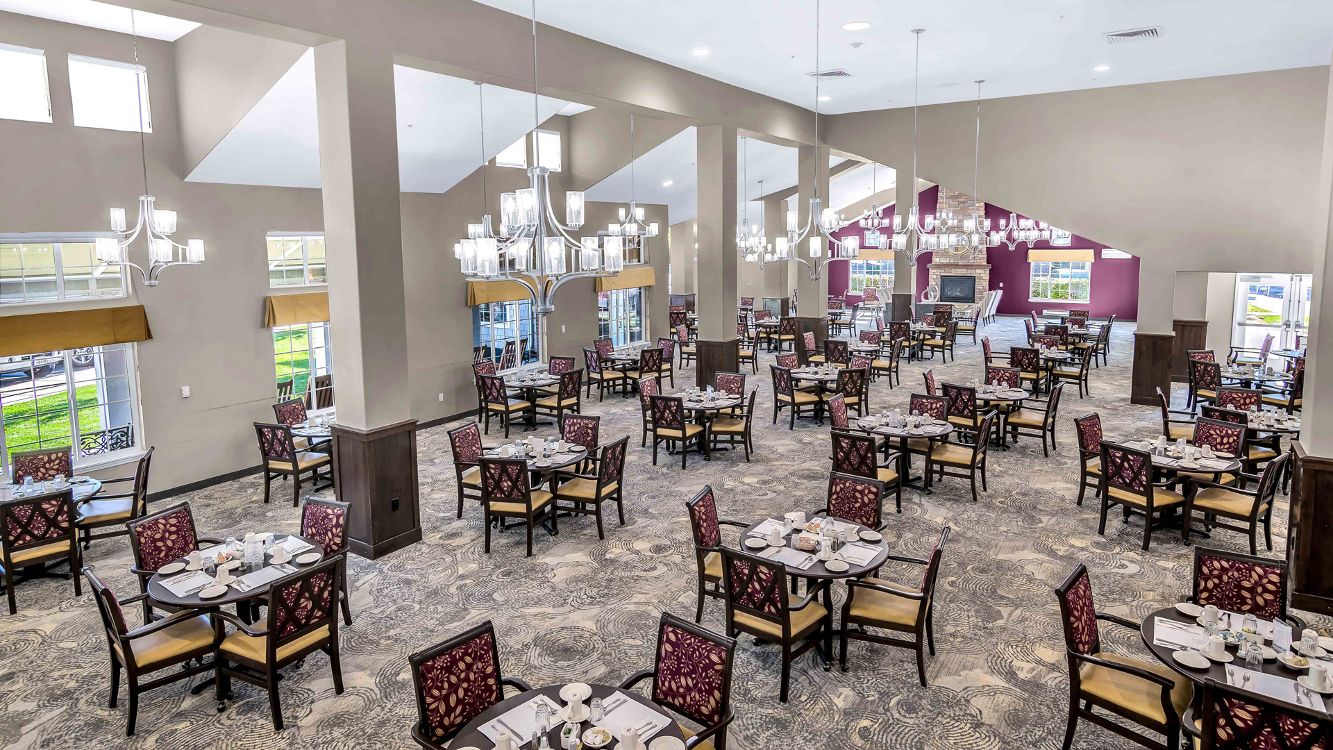 Spacious dining hall with high ceilings, many tables set with plates and cups, patterned carpet, large windows, and modern chandeliers. The room is brightly lit and ready for guests.