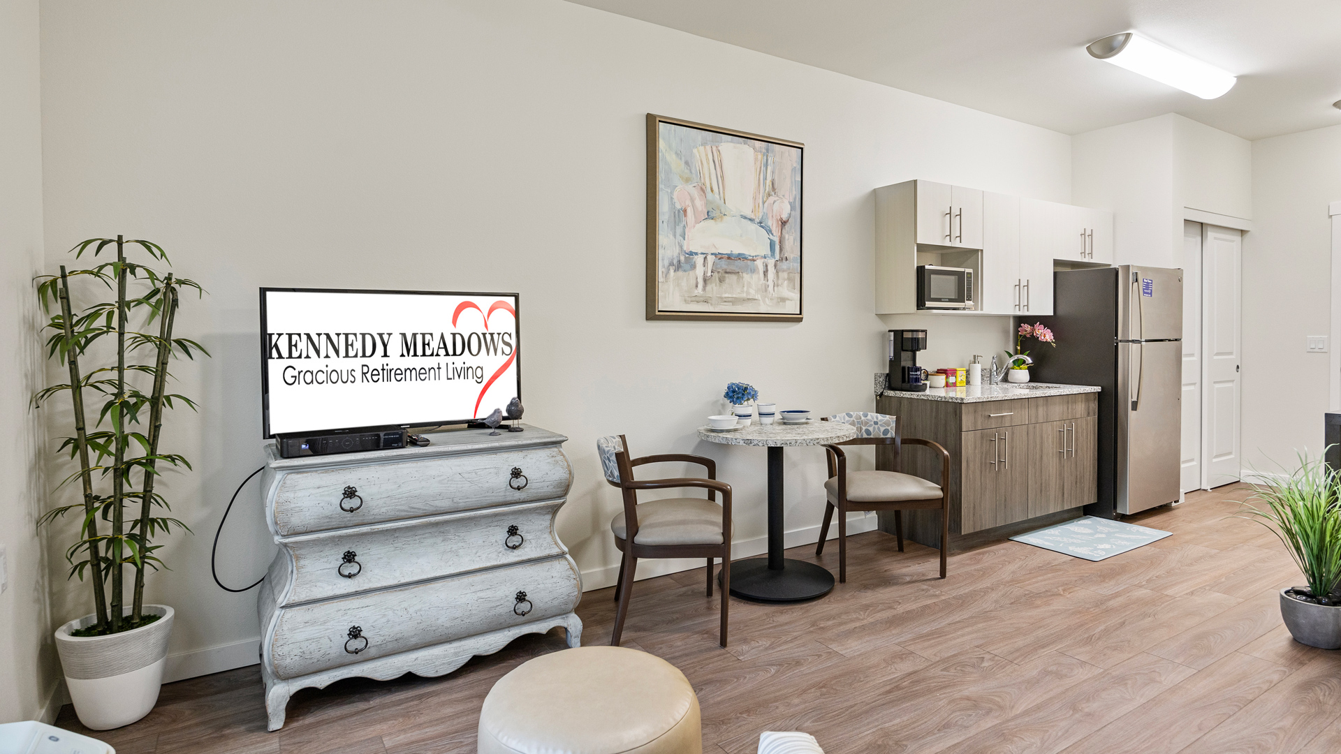 A cozy living area with a TV on a vintage dresser, a small dining table with two chairs, a kitchenette with modern appliances, and a potted plant in the corner. The TV displays "KENNEDY MEADOWS Gracious Retirement Living.