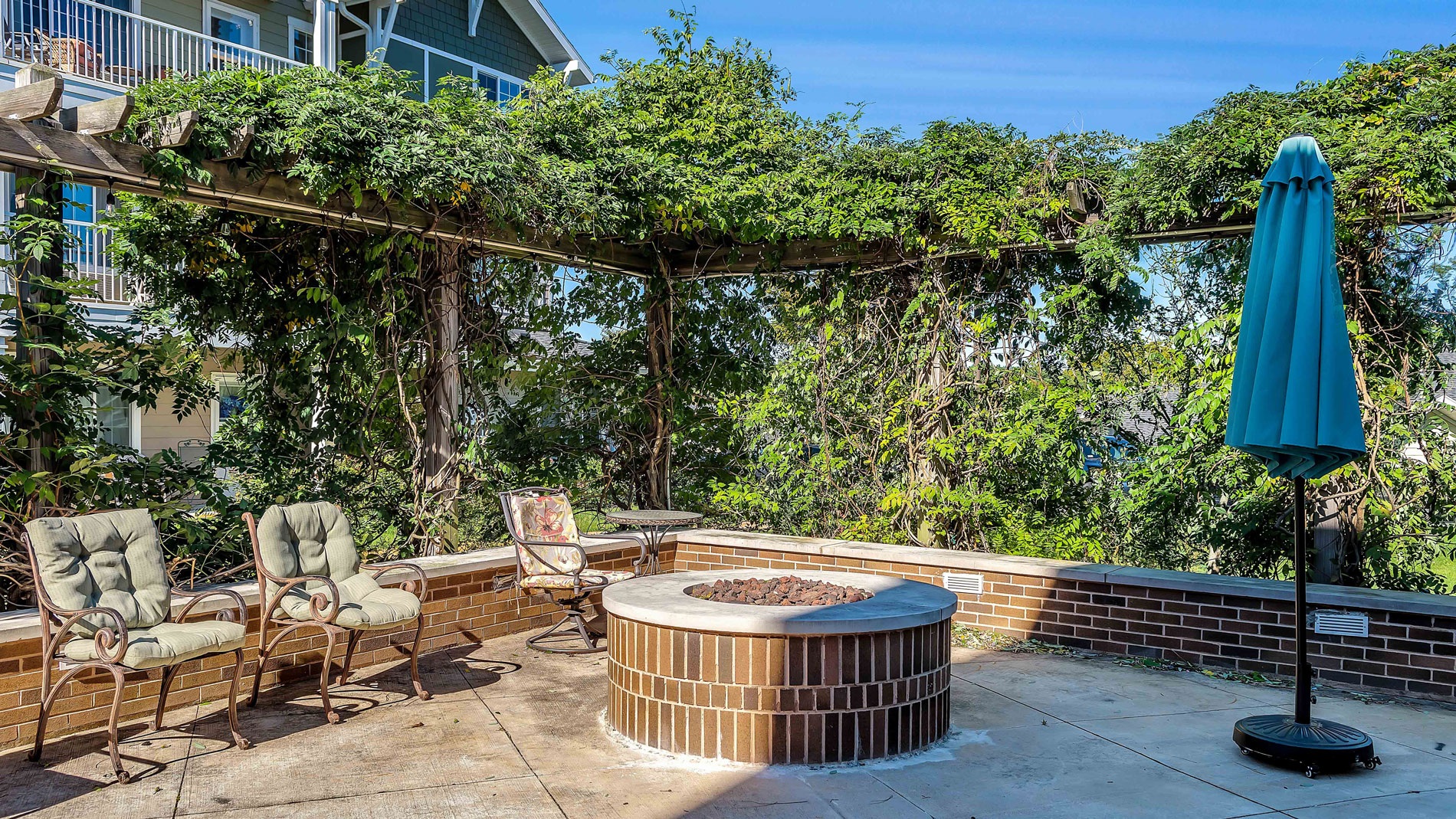 Outdoor patio with cushioned chairs, a small table, a round brick fire pit, and a closed teal umbrella, surrounded by lush green vines on a wooden pergola under a clear blue sky.
