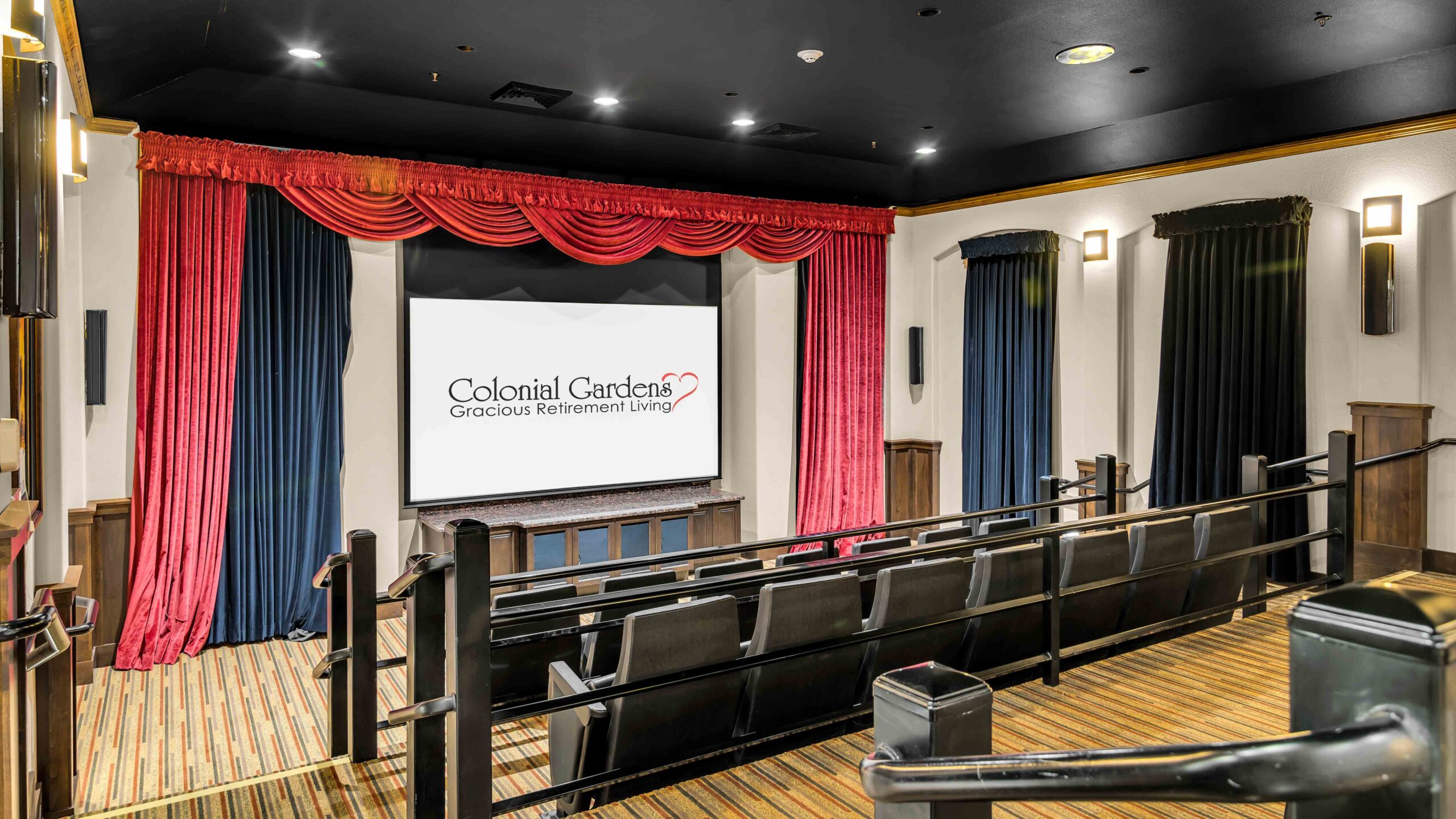 A small theater room with several rows of black seats, a screen displaying "Colonial Gardens Gracious Retirement Living," red stage curtains, wall sconces, and blue drapes on the walls.