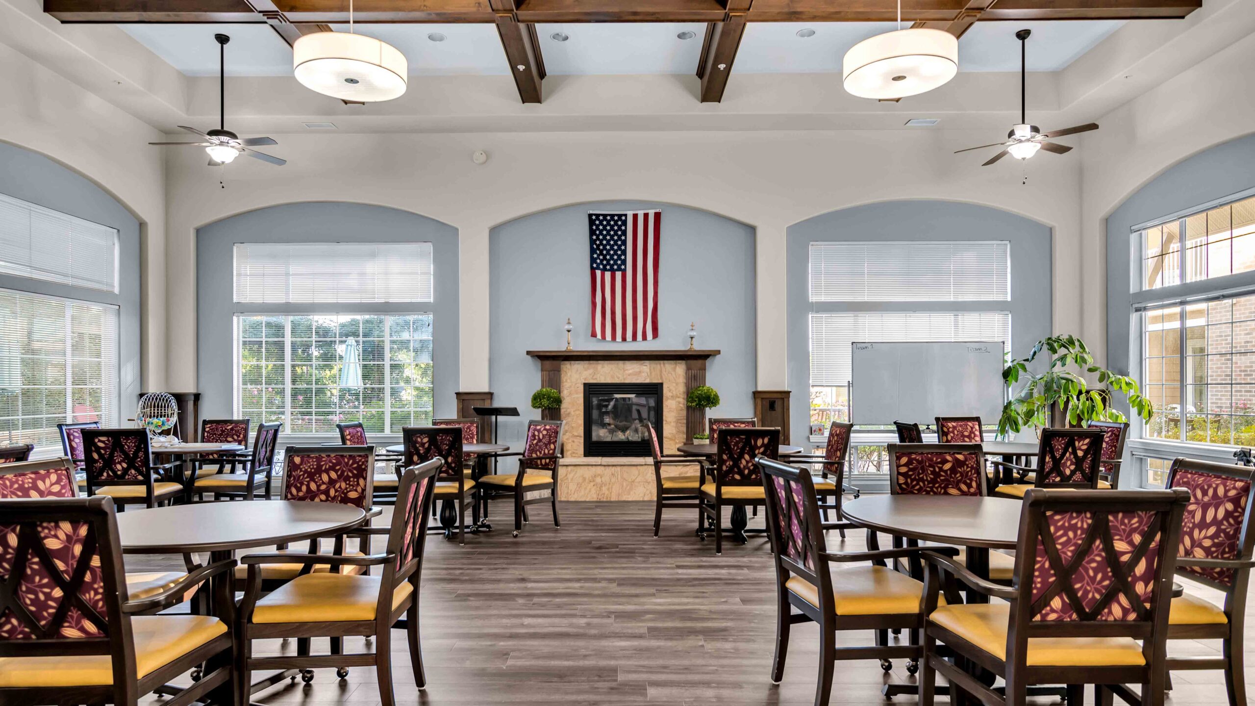 Spacious room with several round tables and chairs, large windows, a fireplace with an American flag above it, ceiling fans, pendant lights, and a whiteboard on the right.