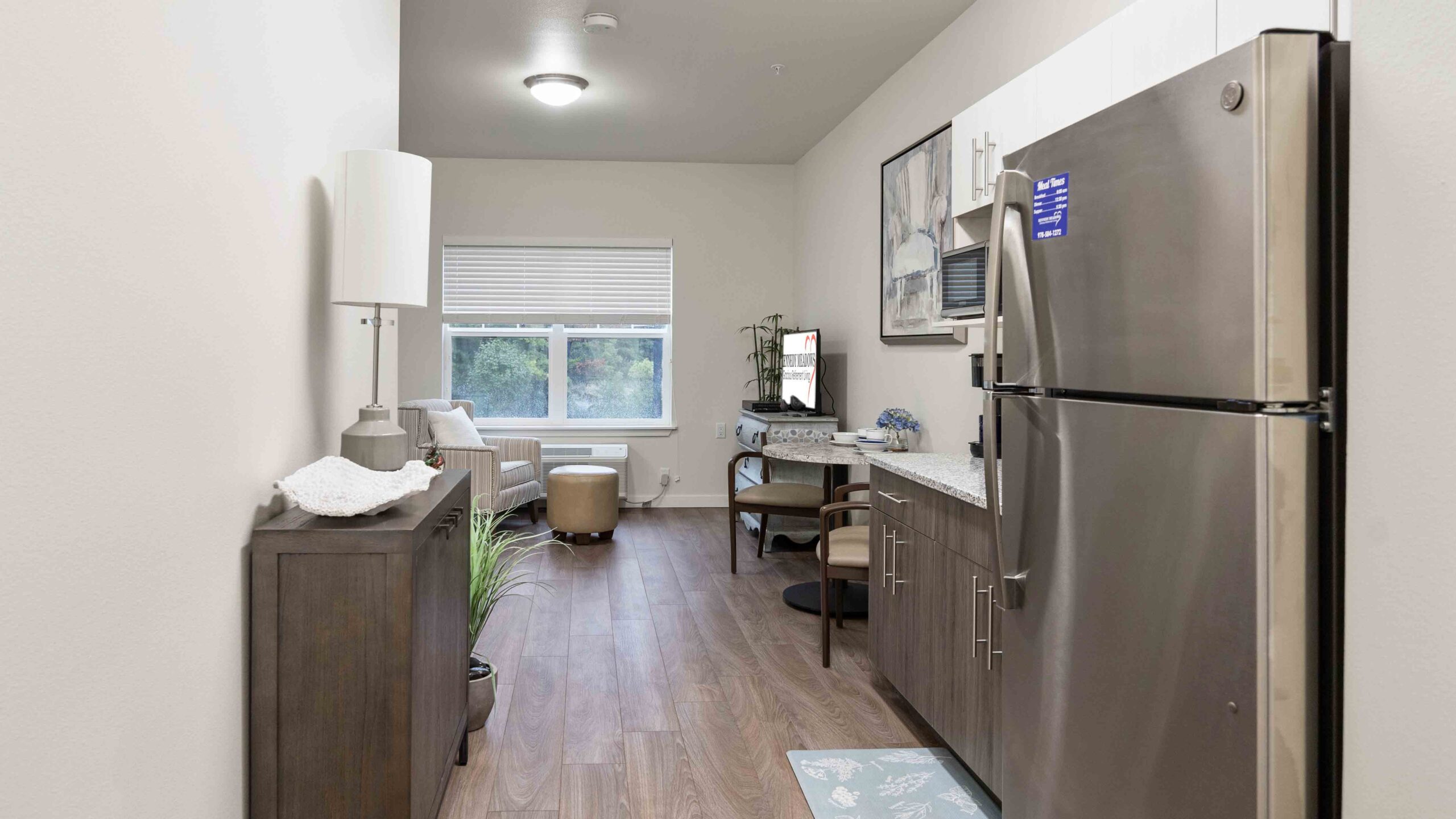 A modern, compact kitchen with stainless steel appliances, white cabinets, and wood flooring leads to a cozy sitting area by a window with two chairs and natural light filling the room.