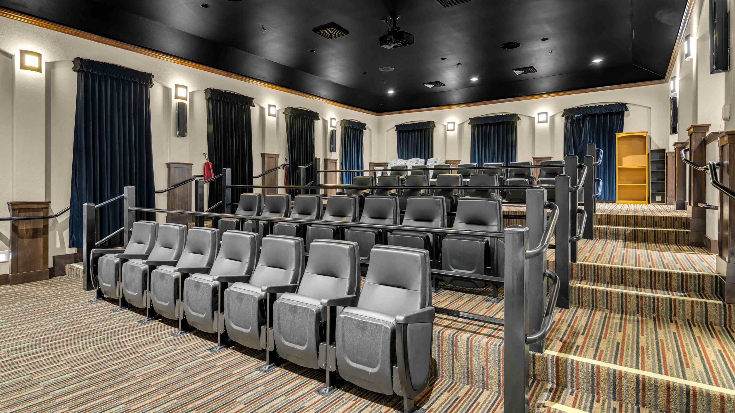 A modern home theater room with tiered rows of black leather seats, striped carpet, wall lights, dark curtains, and a large blank screen at the front.