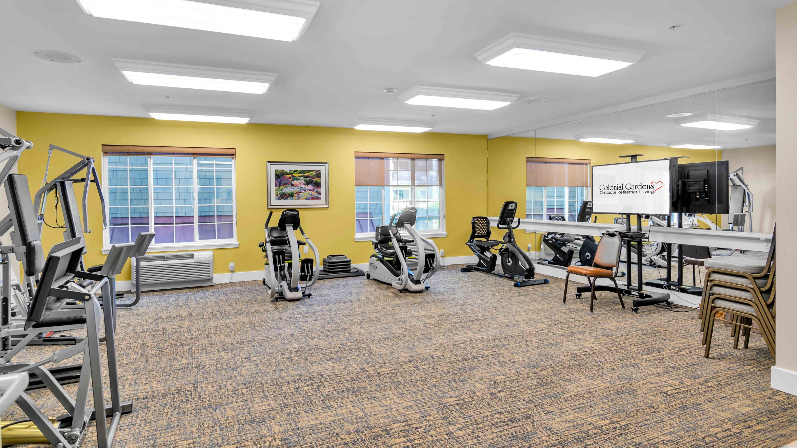 A brightly lit fitness room with exercise equipment, including stationary bikes, a treadmill, resistance machines, and stacked chairs. Large windows, a wall-mounted TV, and framed artwork are visible on the yellow and beige walls.