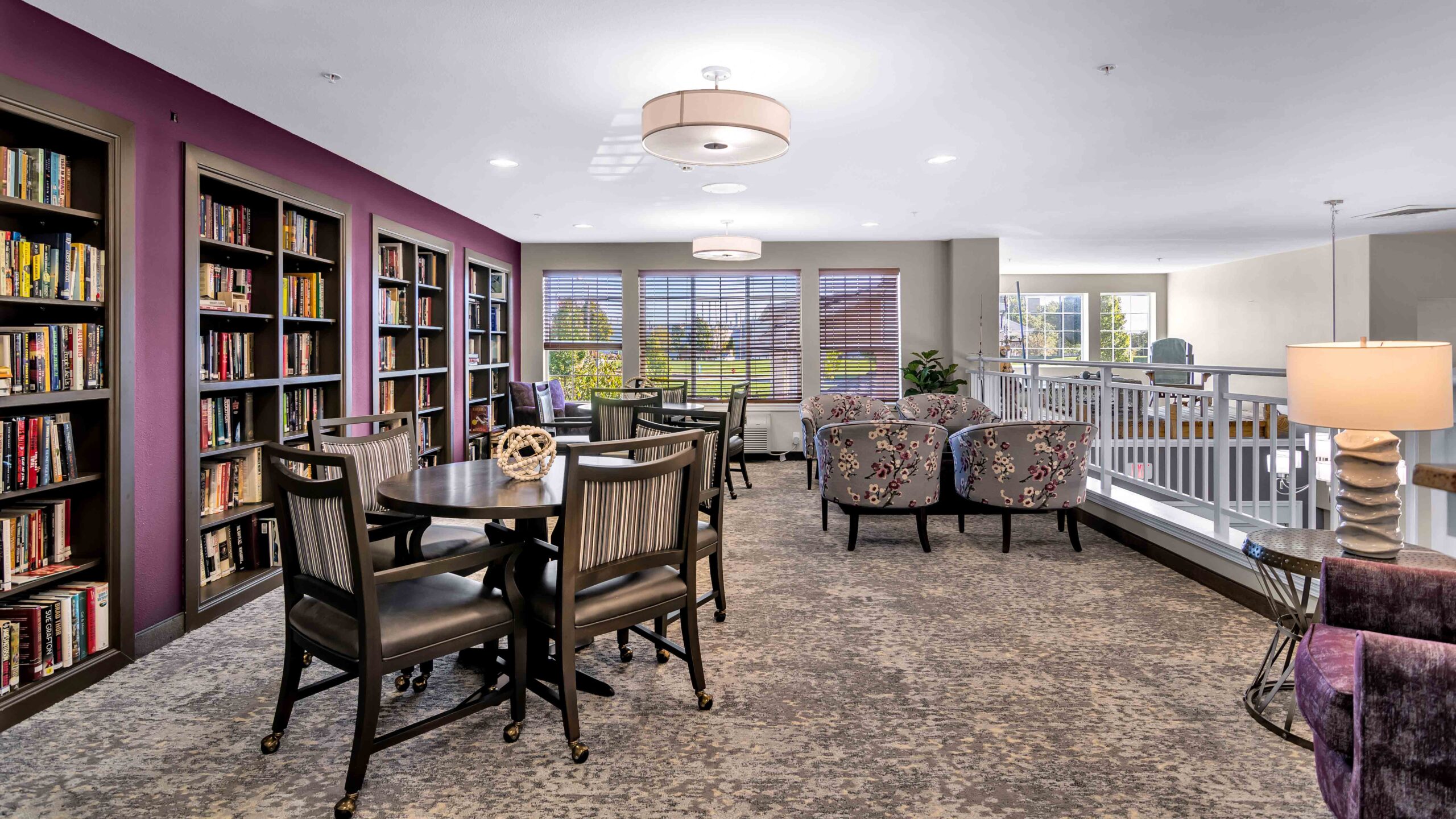 A cozy library lounge with bookshelves, tables and chairs, armchairs by large windows, and soft lighting. The space features purple accents and overlooks a bright, open area with railing.