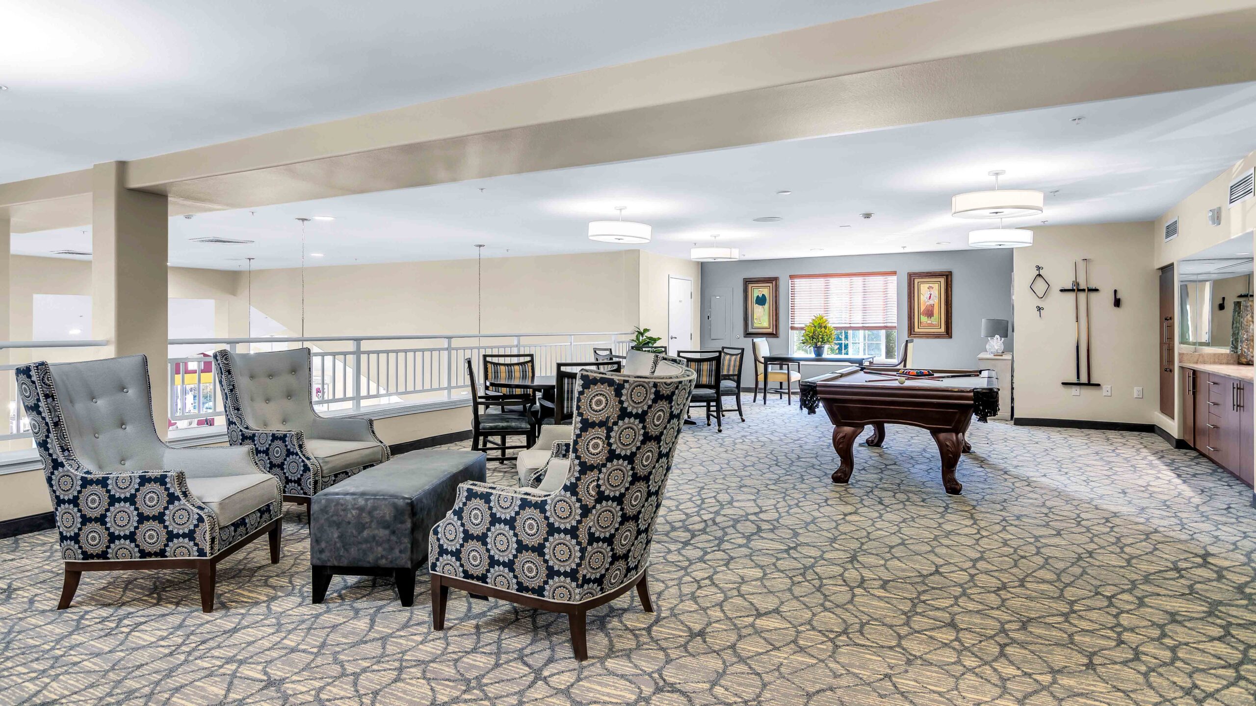 A spacious, well-lit lounge area with patterned armchairs, a cushioned bench, small tables and chairs, a pool table, artwork on the walls, and a large window with blinds letting in natural light.