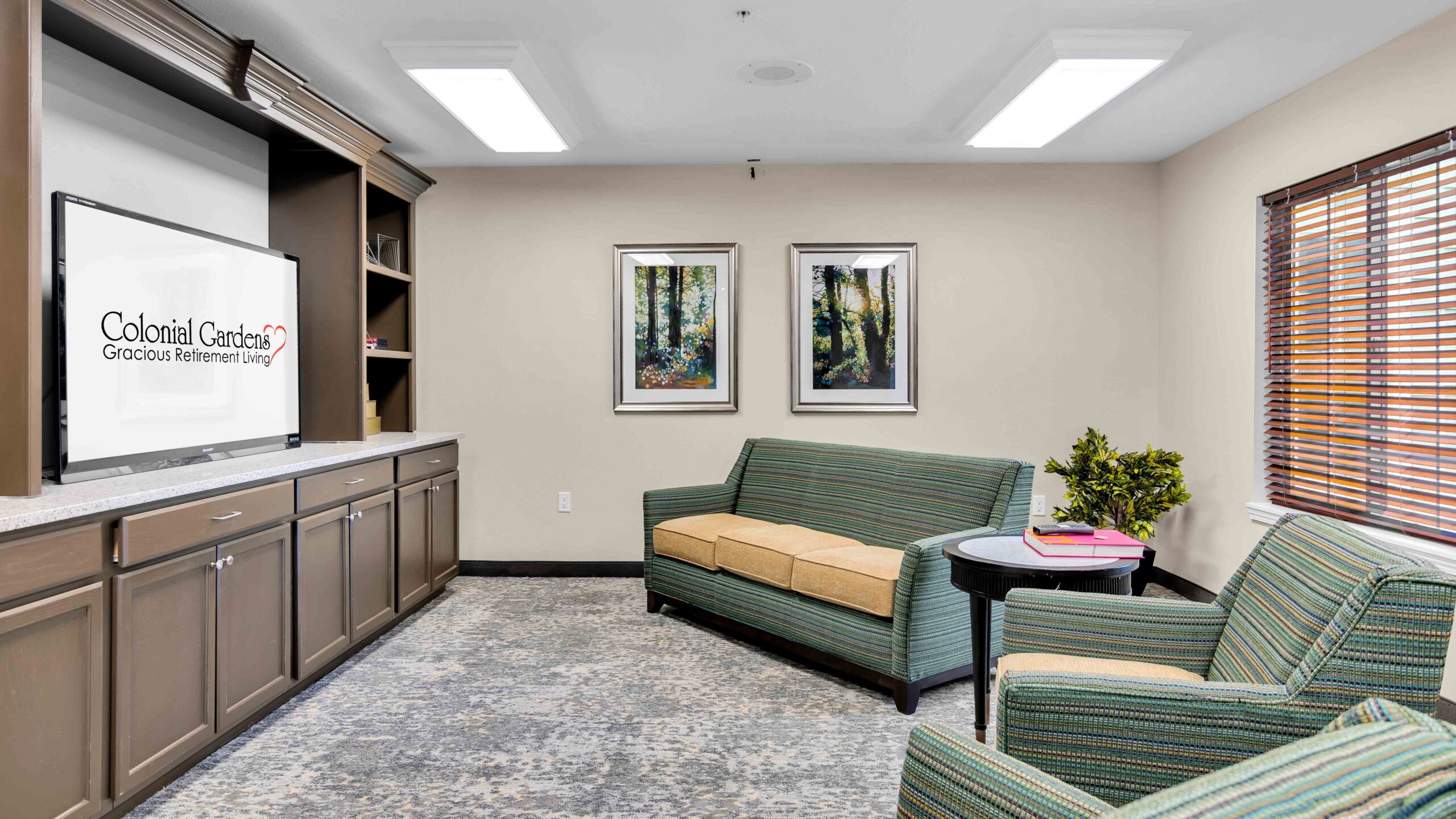 A cozy lounge area with a green sofa, armchair, small table, and a TV displaying "Colonial Gardens Gracious Retirement Living." Two framed nature prints hang on the beige wall, and a window has wooden blinds.