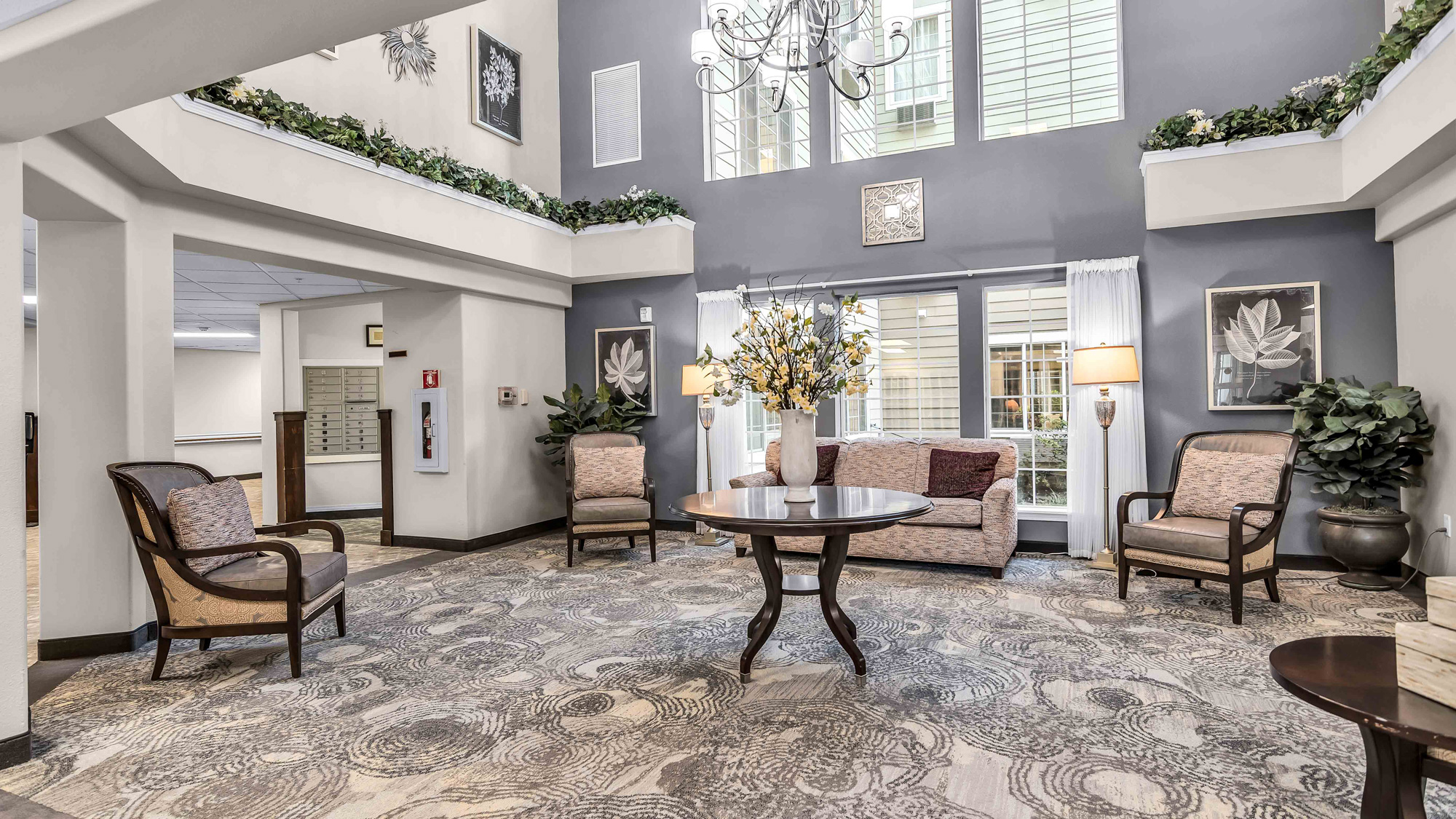 A spacious, elegant lobby with a round table and flower arrangement in the center, surrounded by armchairs and a sofa. Large windows, plants, framed art, and neutral tones create a welcoming atmosphere.