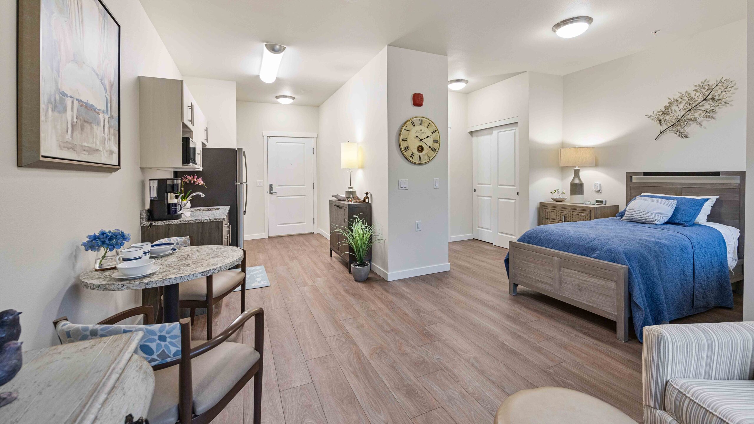 A modern studio apartment with wood floors, a small kitchen, dining area, and a bed with blue bedding. Light walls, simple decor, and soft lighting create a cozy, inviting atmosphere.
