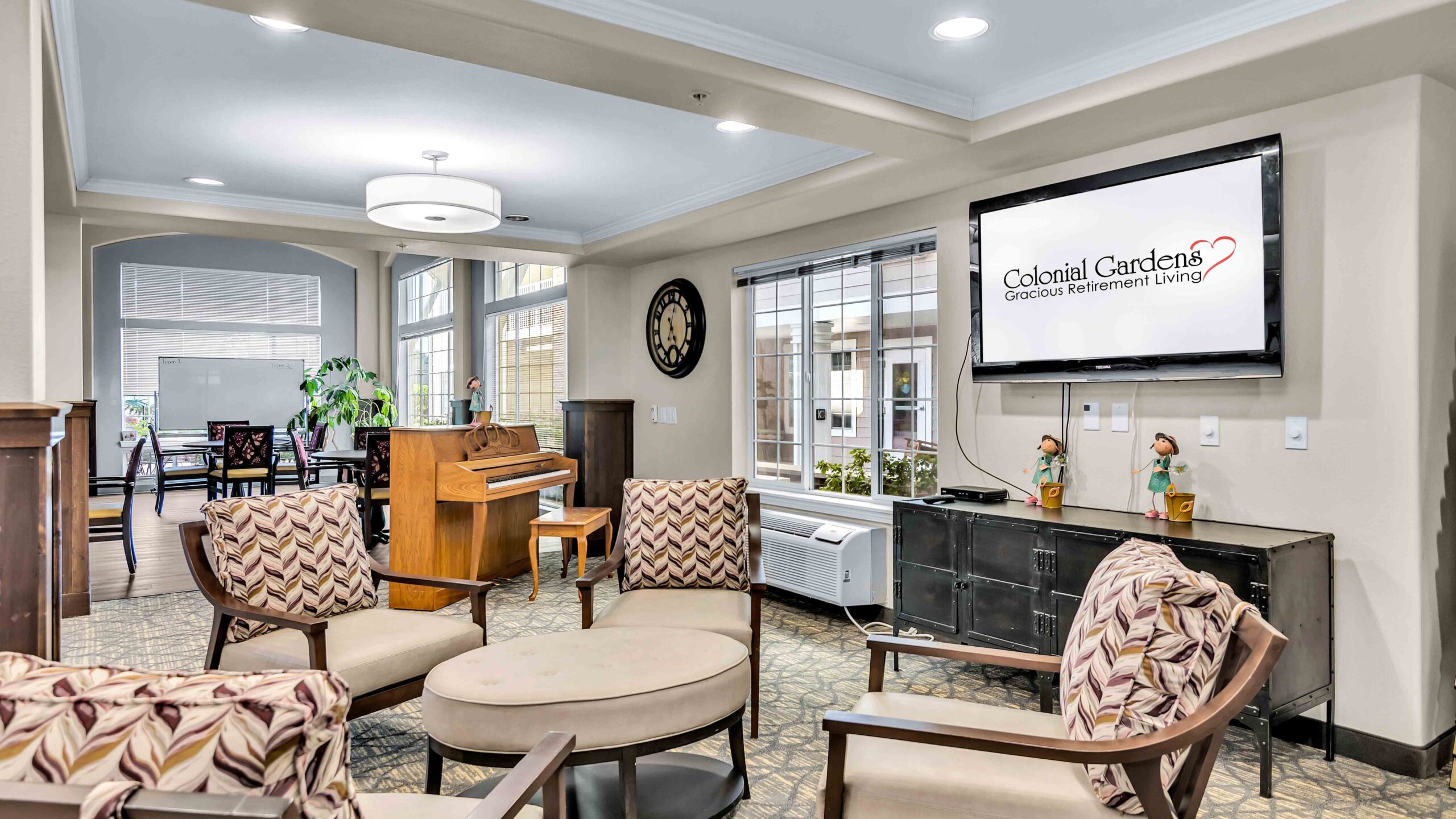 A cozy retirement living room with patterned chairs around a circular table, a piano, flat-screen TV displaying "Colonial Gardens Gracious Retirement Living," and large windows letting in natural light.
