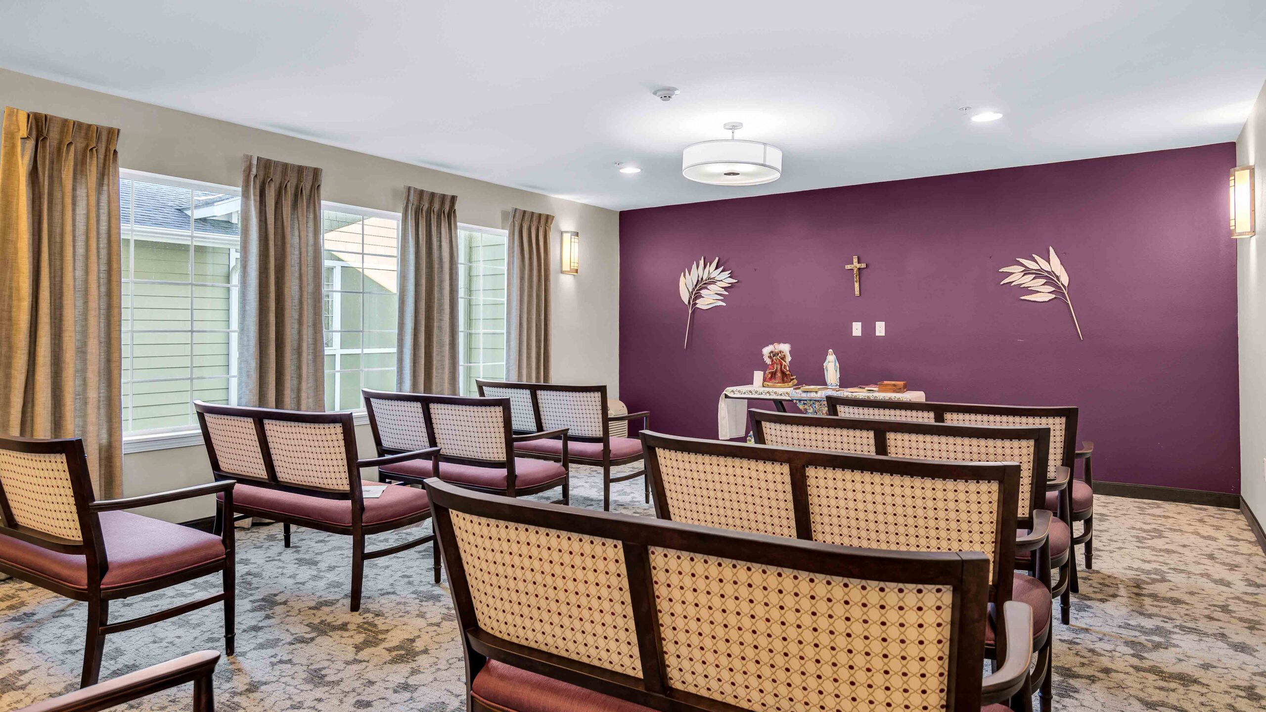 Small chapel room with rows of chairs facing a purple accent wall, featuring a crucifix, two religious statues, and palm leaf decorations. Large windows with beige curtains let in natural light.