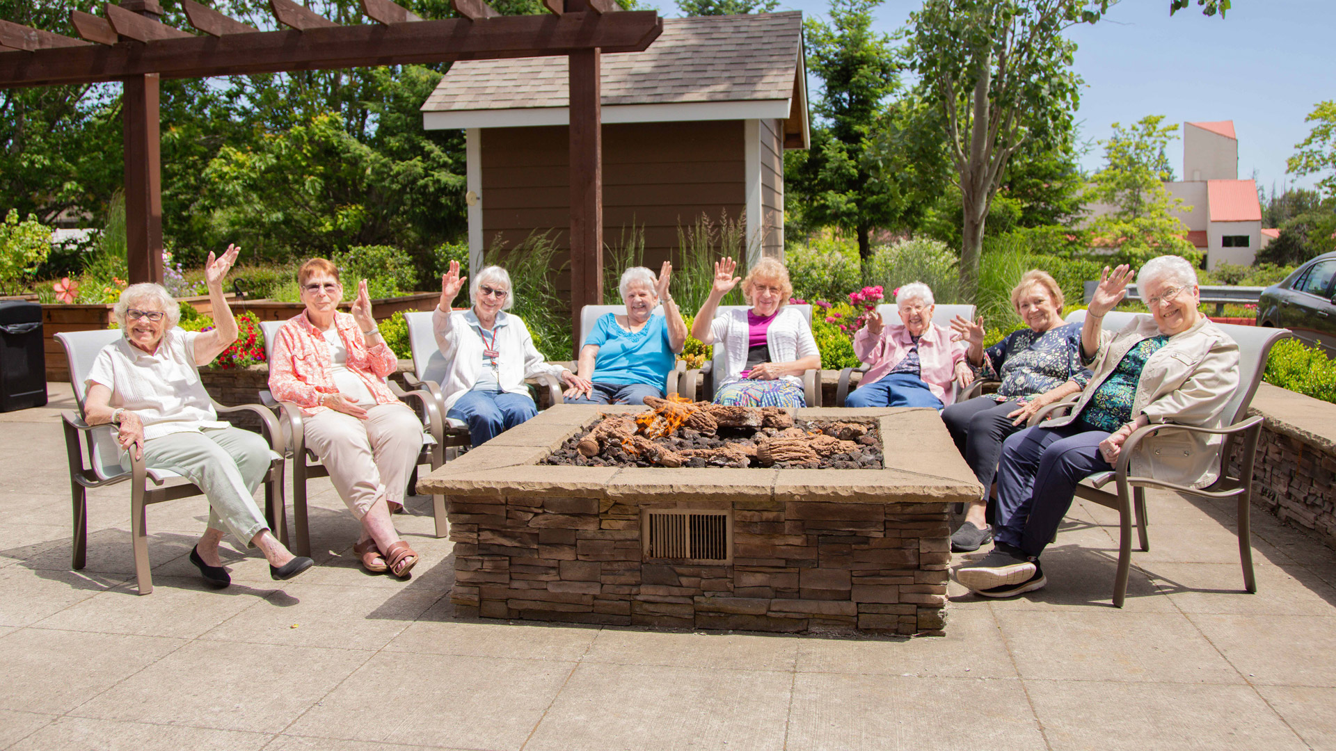 A group of eight elderly women sit in a circle around an outdoor fire pit, smiling and waving at the camera on a sunny day, with greenery and buildings in the background.