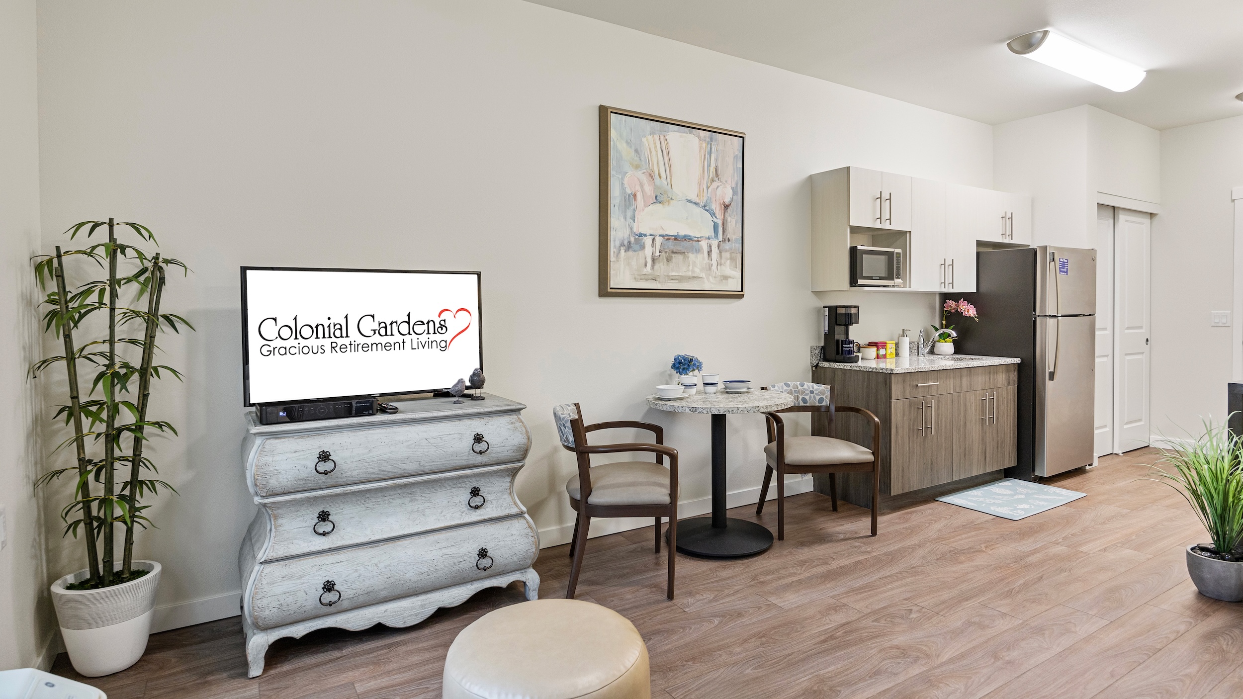 A cozy retirement living space with a small kitchen, dining table with two chairs, TV on a dresser, a painting on the wall, potted plants, and wood flooring. The TV displays “Colonial Gardens Gracious Retirement Living.”.