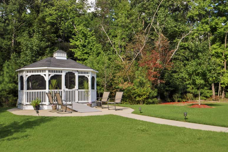 A white wooden gazebo with a gray roof stands on a concrete slab in a lush green garden. Two lawn chairs are placed on the slab next to the gazebo. A narrow concrete path curves through the neatly trimmed grass, leading towards the gazebo. Tall trees form a dense backdrop.