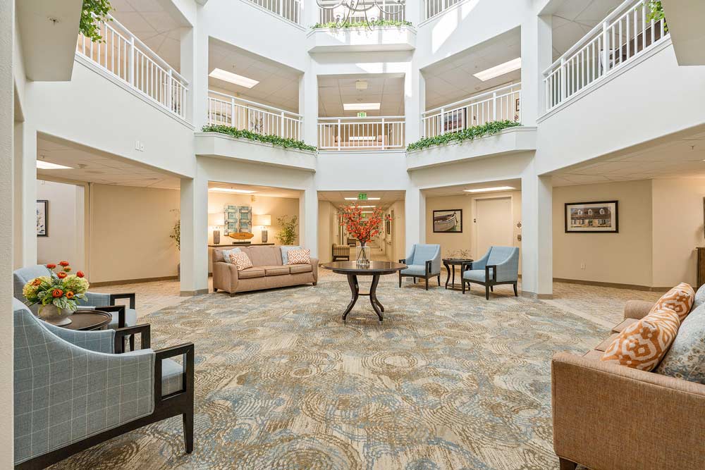 A spacious, light-filled atrium lobby with high ceilings, surrounded by two floors of white railings. There are seating areas with beige sofas, blue chairs, and tables adorned with floral arrangements. The floor is carpeted in a patterned, neutral-toned design.