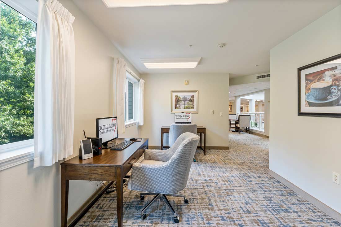 A bright, carpeted room with two wooden desks, each equipped with a computer and gray office chair. A window on the left provides natural light, and framed art hangs on the walls. An open doorway at the back reveals more of the space beyond.