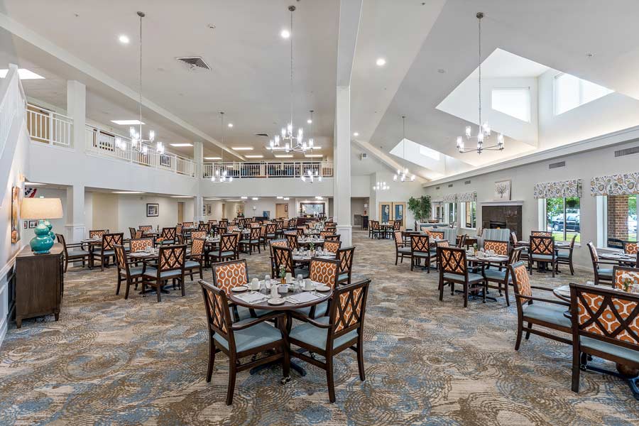 A spacious dining hall with high ceilings and chandeliers, featuring multiple tables and chairs arranged neatly. Bright lighting fills the room, and windows on one side offer natural light. The room has a mix of modern and classic decor elements.