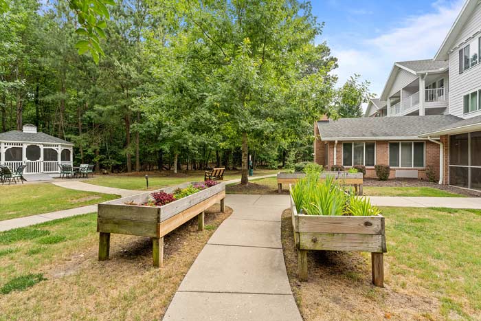 A well-kept outdoor area with a paved walkway surrounded by raised garden beds filled with various plants. To the left stands a white gazebo with seating, while a two-story building is visible to the right. Trees and greenery fill the background.