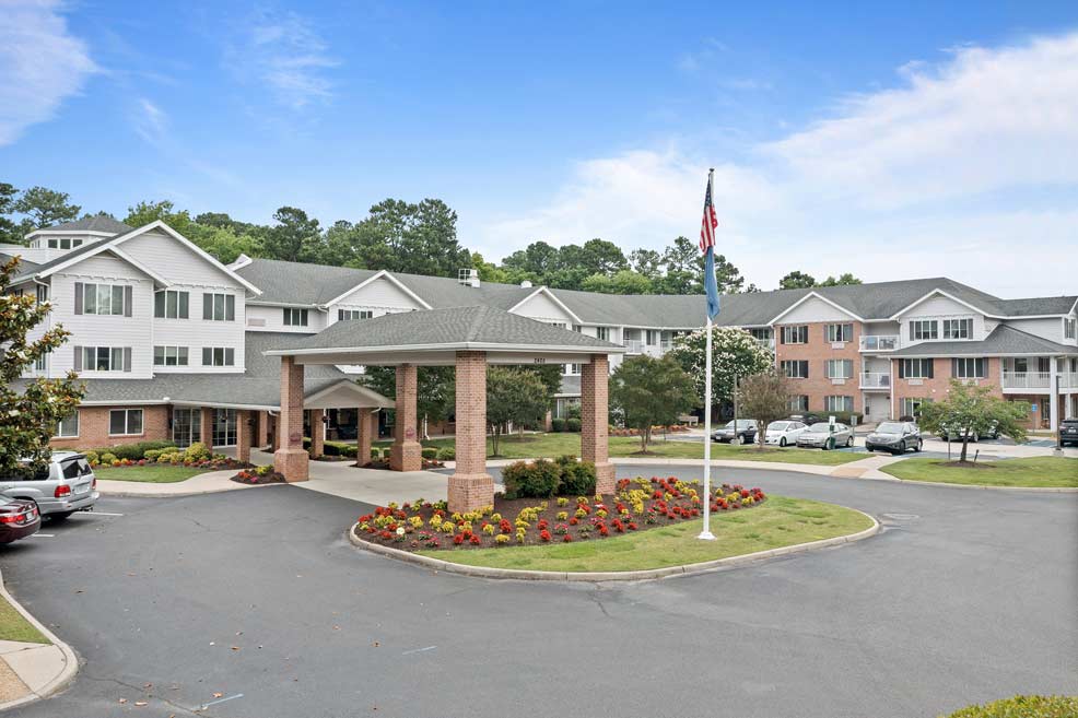 A large, multi-story residential building with a prominent entrance featuring four pillars and a covered driveway. A well-maintained garden with colorful flowers and an American flag on a pole can be seen in front. Several cars are parked around the building.