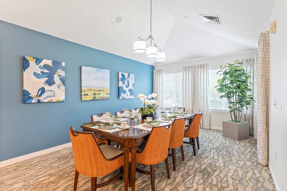 A dining room features a rectangular wooden table set with plates and glasses, surrounded by eight chairs with orange cushions. The walls are painted blue and decorated with three abstract art pieces. Large windows with sheer curtains and a potted plant complete the space.