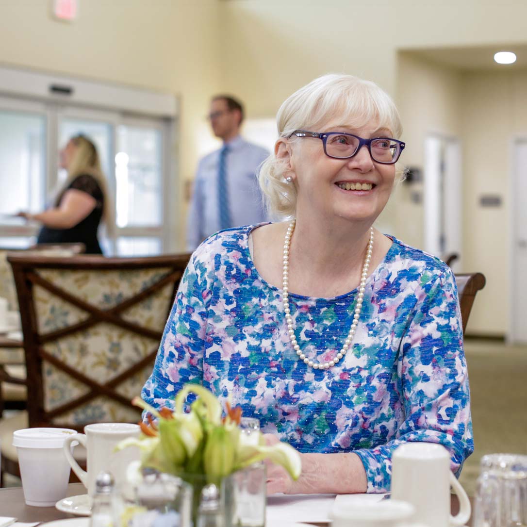 An elderly woman with white hair and glasses, wearing a blue floral top and a pearl necklace, smiles while sitting at a table in a bright room. There are cups and a vase of flowers on the table. Two people are in the blurred background.