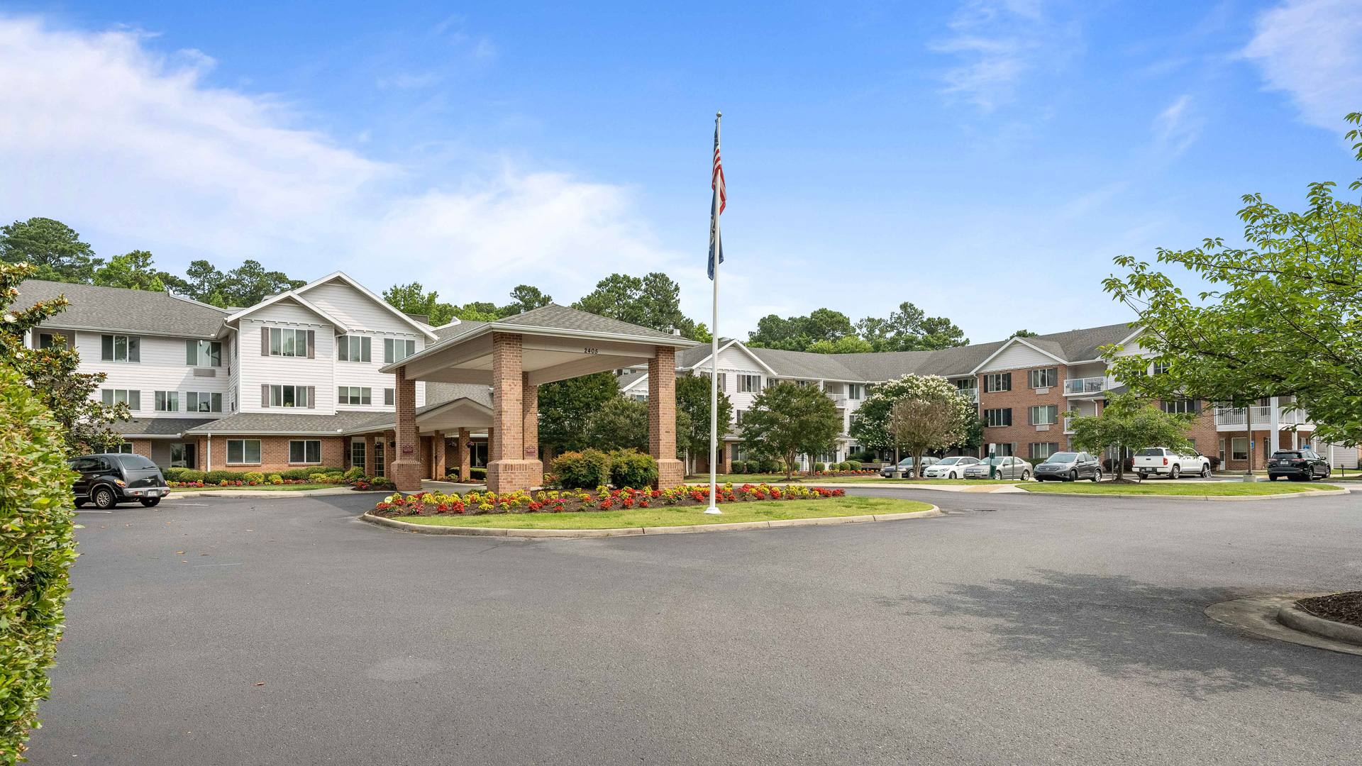 A large, three-story residential building with white and brick exterior sits behind a circular driveway, landscaped flower beds, and a flagpole, under a blue sky with scattered clouds.