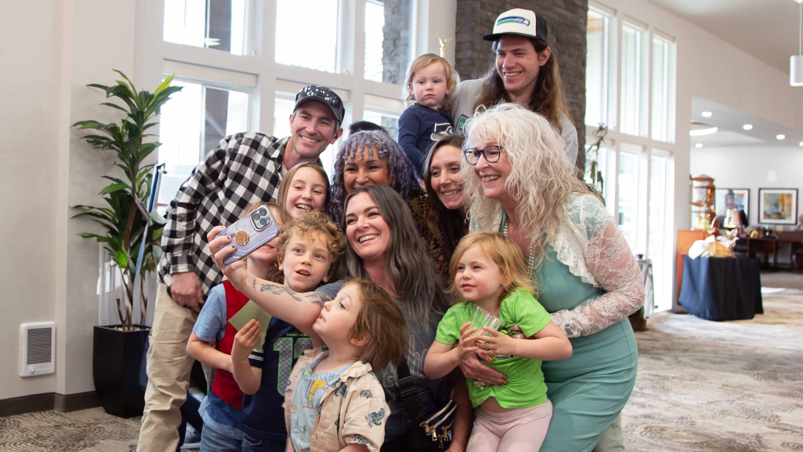 A group of adults and children gather closely together, smiling for a selfie in a bright, modern indoor space with large windows and plants in the background.