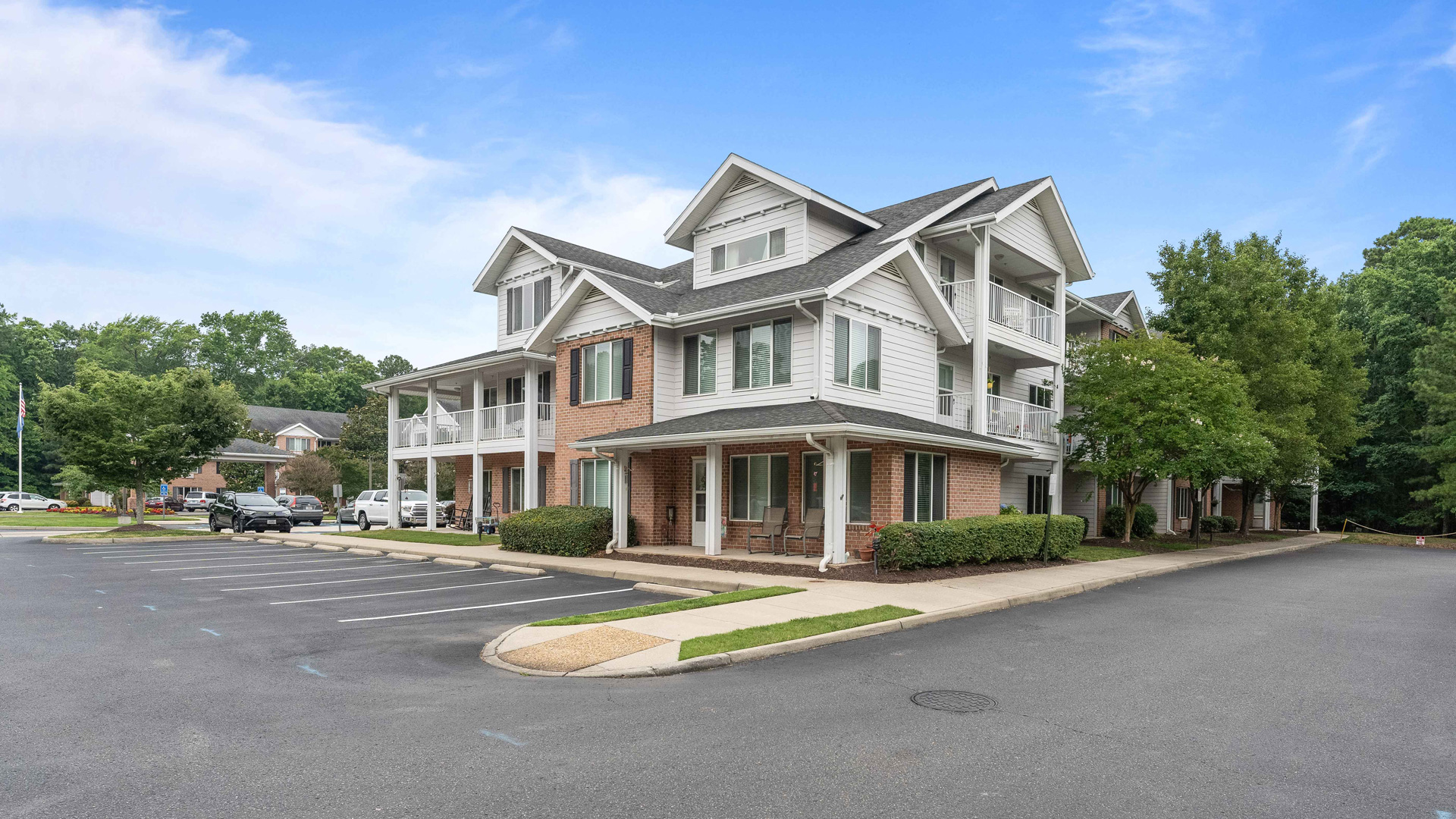 A two-story apartment building with white trim, brick and siding exterior, multiple balconies, and a surrounding parking lot with a few cars, set on a clear day with green trees in the background.