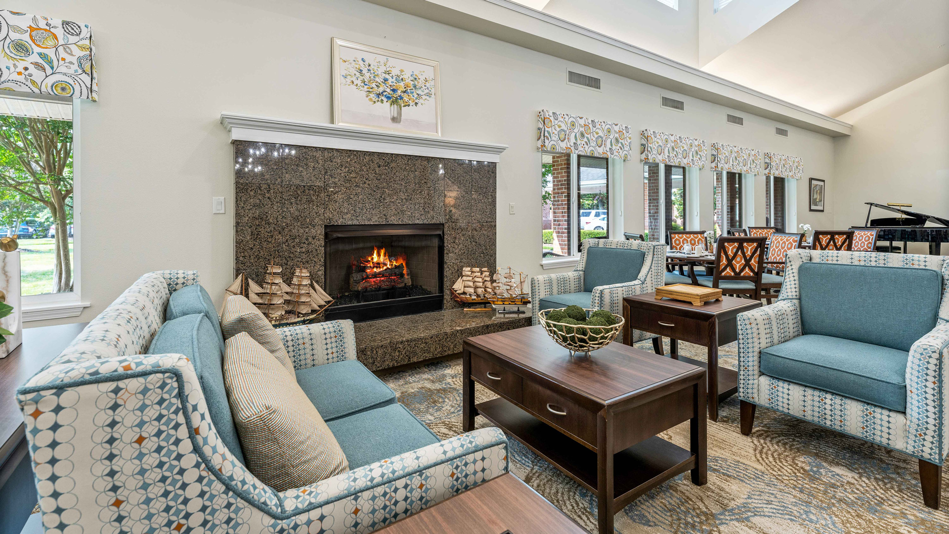 A cozy living room with blue armchairs, a dark wooden coffee table, a fireplace with a fire burning, ship model decorations, and large windows letting in natural light.