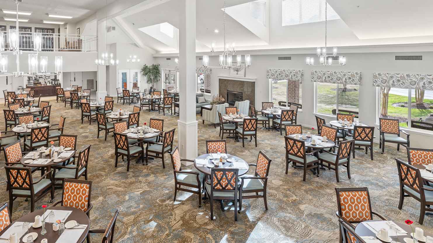 A spacious, well-lit dining room with many round tables set for meals, surrounded by patterned chairs. Large windows, chandeliers, and neutral decor create an inviting, elegant atmosphere.