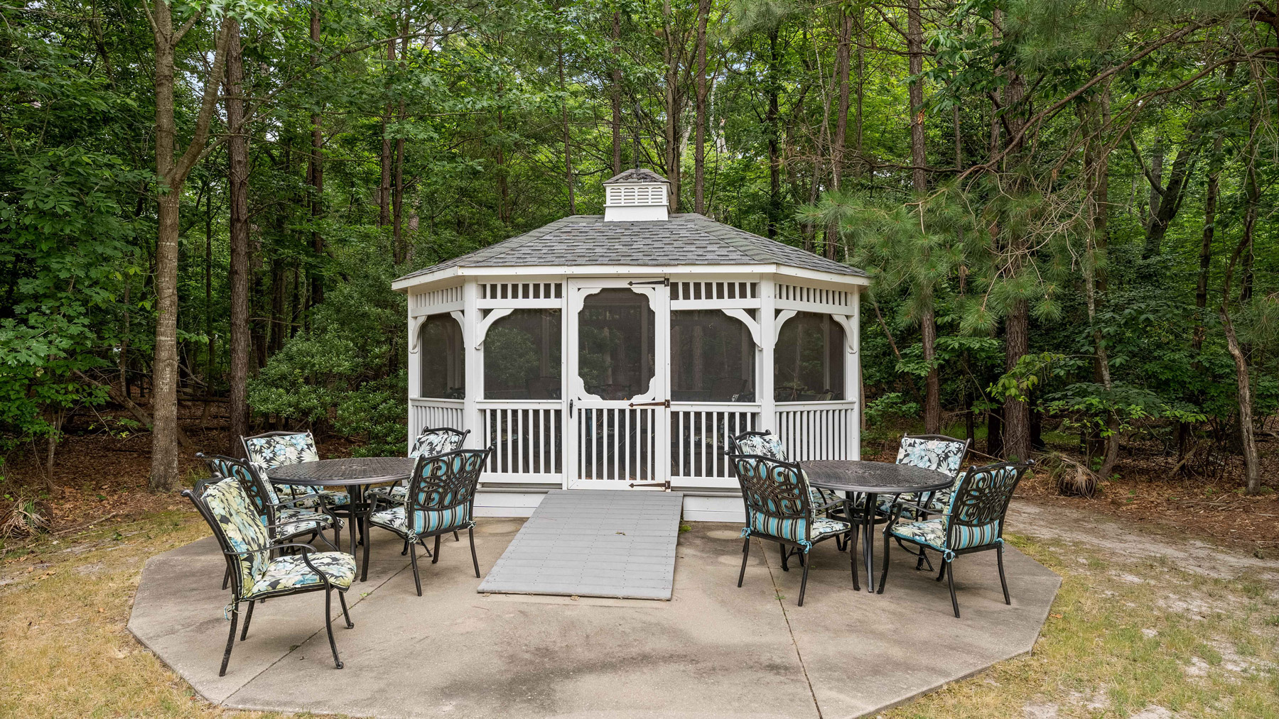 A white, screened gazebo sits on a concrete pad surrounded by trees, with two round tables and eight floral-cushioned chairs arranged outdoors around it.