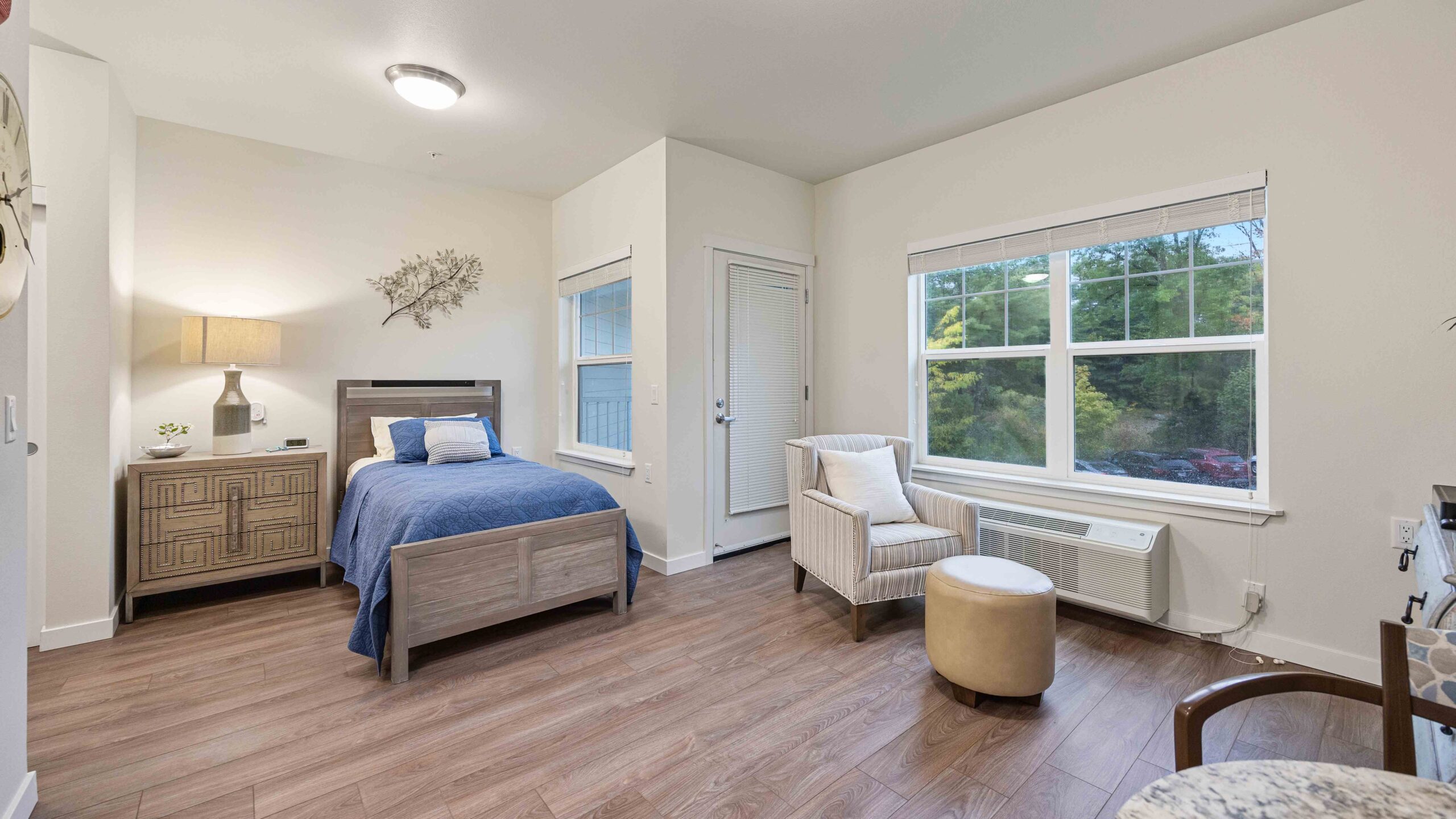 A cozy, well-lit bedroom with a single bed, wooden nightstand, lamp, padded chair, ottoman, and large window overlooking greenery. The floor is wood, and decor is minimal with neutral tones.
