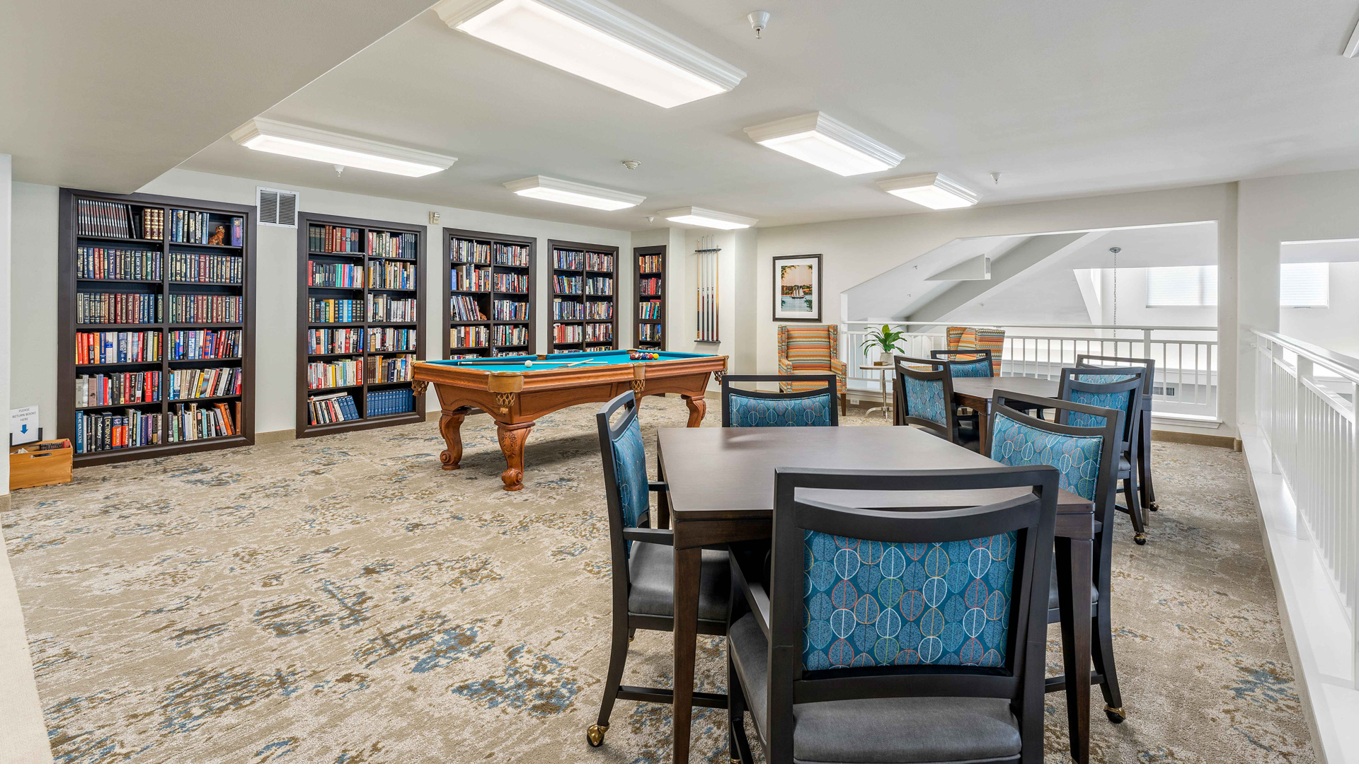 A spacious library room with bookshelves, a pool table, and a table with blue-patterned chairs, all set on a beige carpet under bright ceiling lights.
