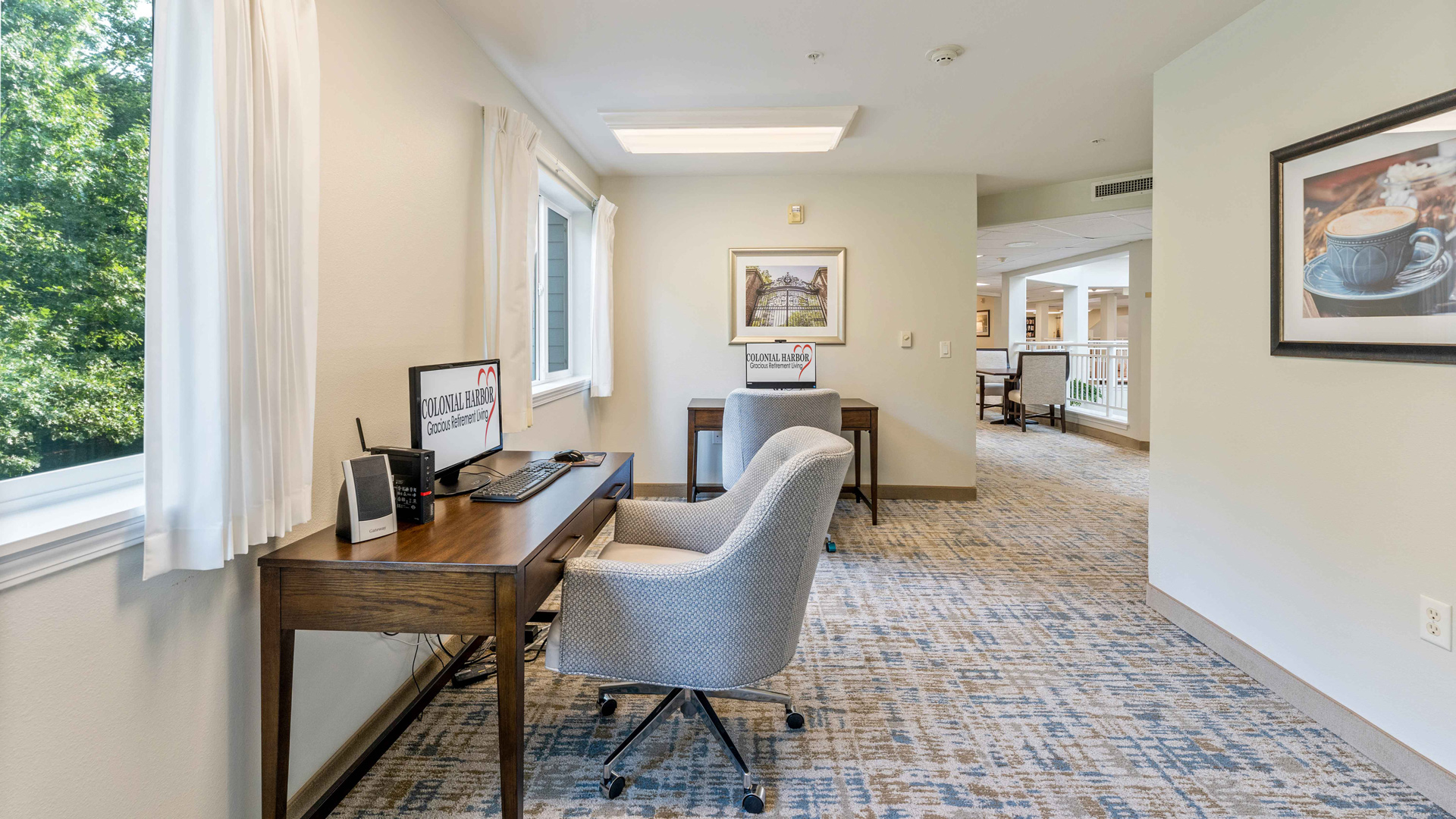 A bright room with two desks, each with a computer and a gray cushioned chair, sits beside a large window with white curtains and overlooks a cozy hallway with framed artwork and carpeted floors.