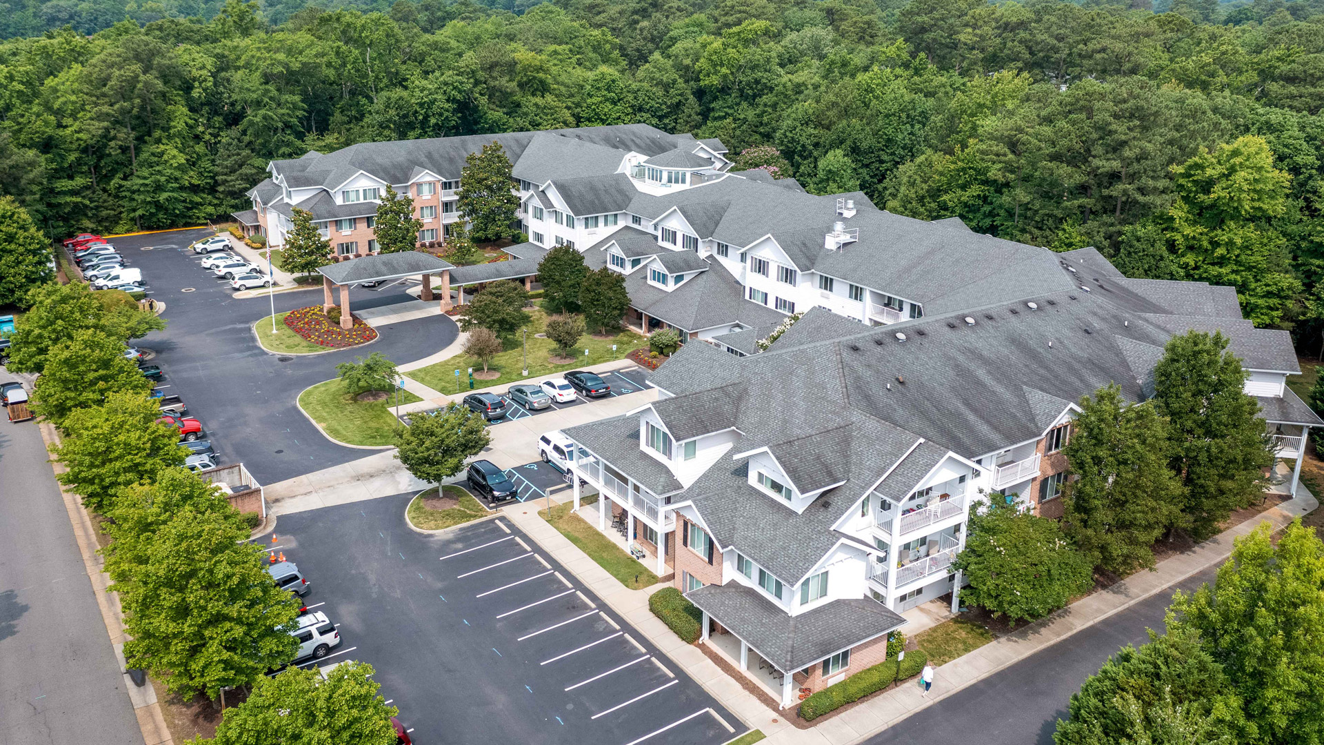 Aerial view of a large residential complex with multiple connected buildings, parking lots with cars, landscaped greenery, and surrounded by dense trees.