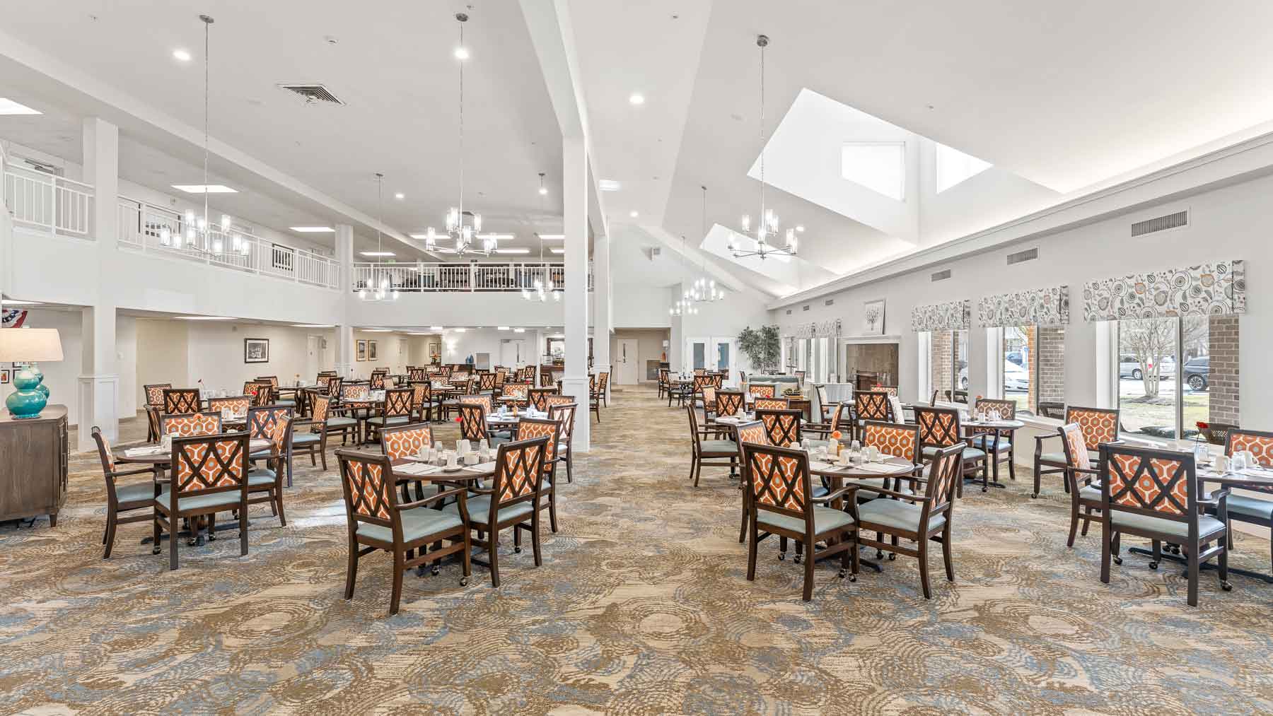 Spacious, well-lit dining hall with many wooden tables and chairs, set for meals. Large windows on the right let in natural light. Stylish chandeliers hang from the high ceiling, and patterned carpet covers the floor.