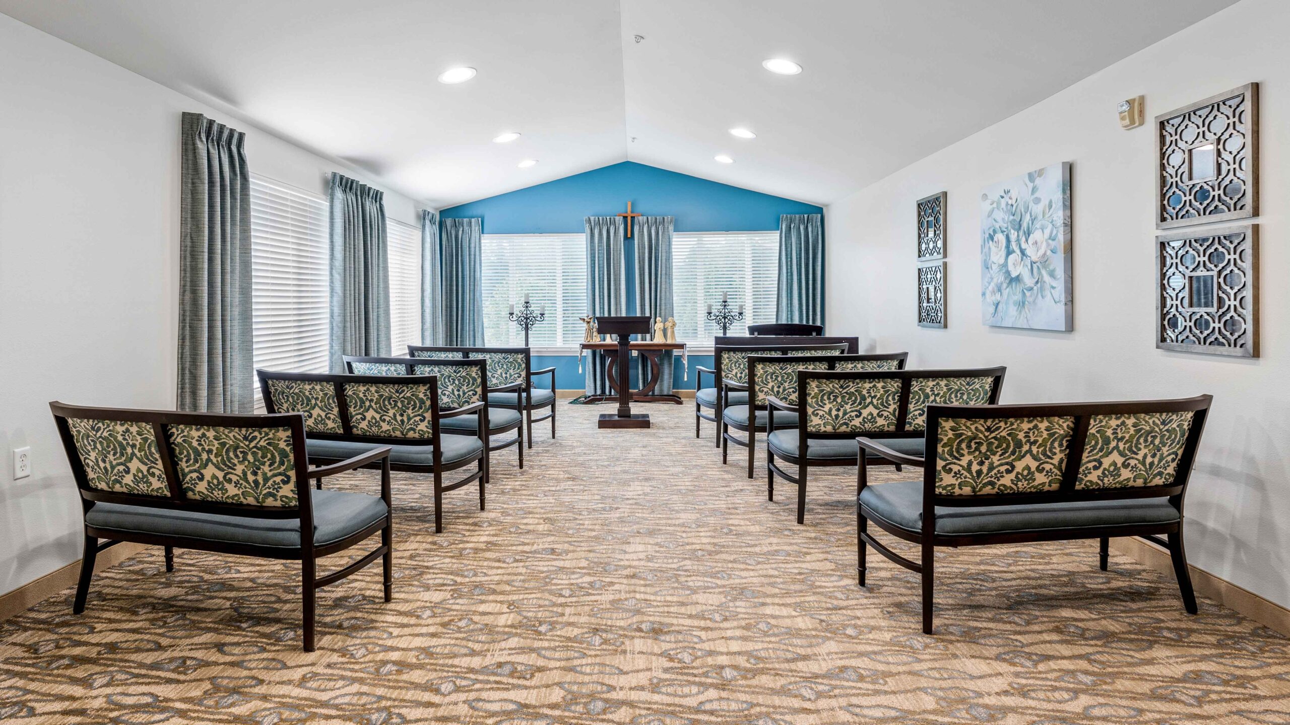 A small chapel with patterned chairs arranged in rows facing a wooden altar. The room has large windows with blue curtains, floral artwork on the walls, and neutral-toned carpet.