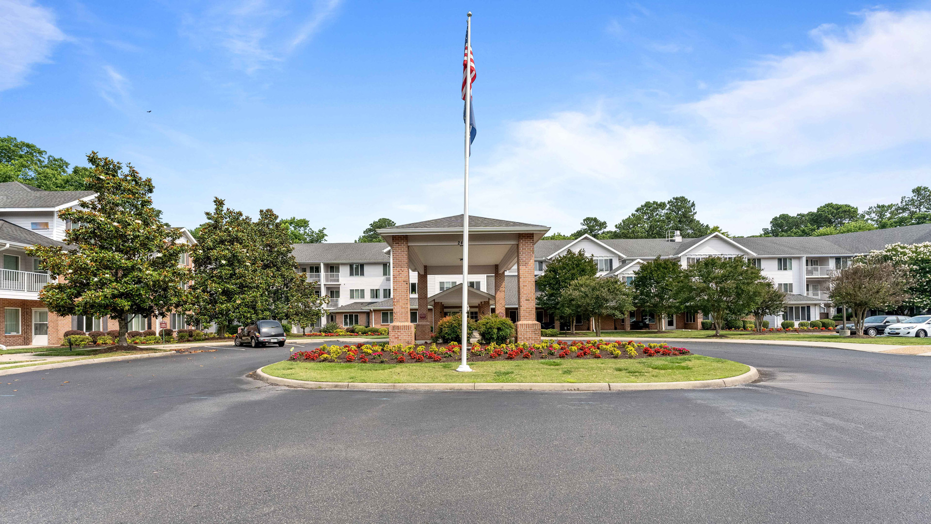 A circular driveway surrounds a landscaped flower bed and flagpole in front of a large, multi-story brick building with balconies and white trim, under a blue sky with scattered clouds.