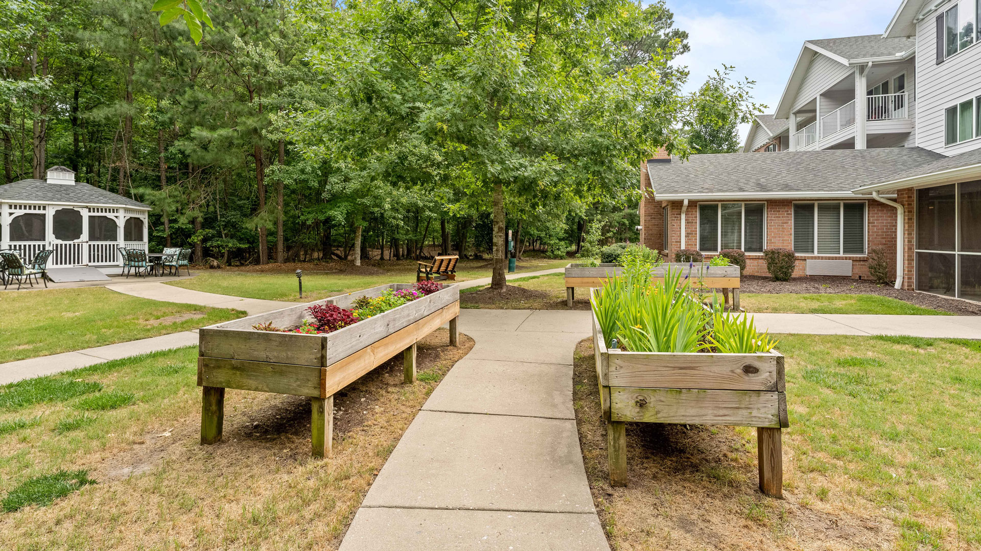 A paved pathway curves through a garden area with raised wooden planters, trees, and a gazebo on the left. The path is surrounded by grass and leads to a building on the right with white railings and windows.