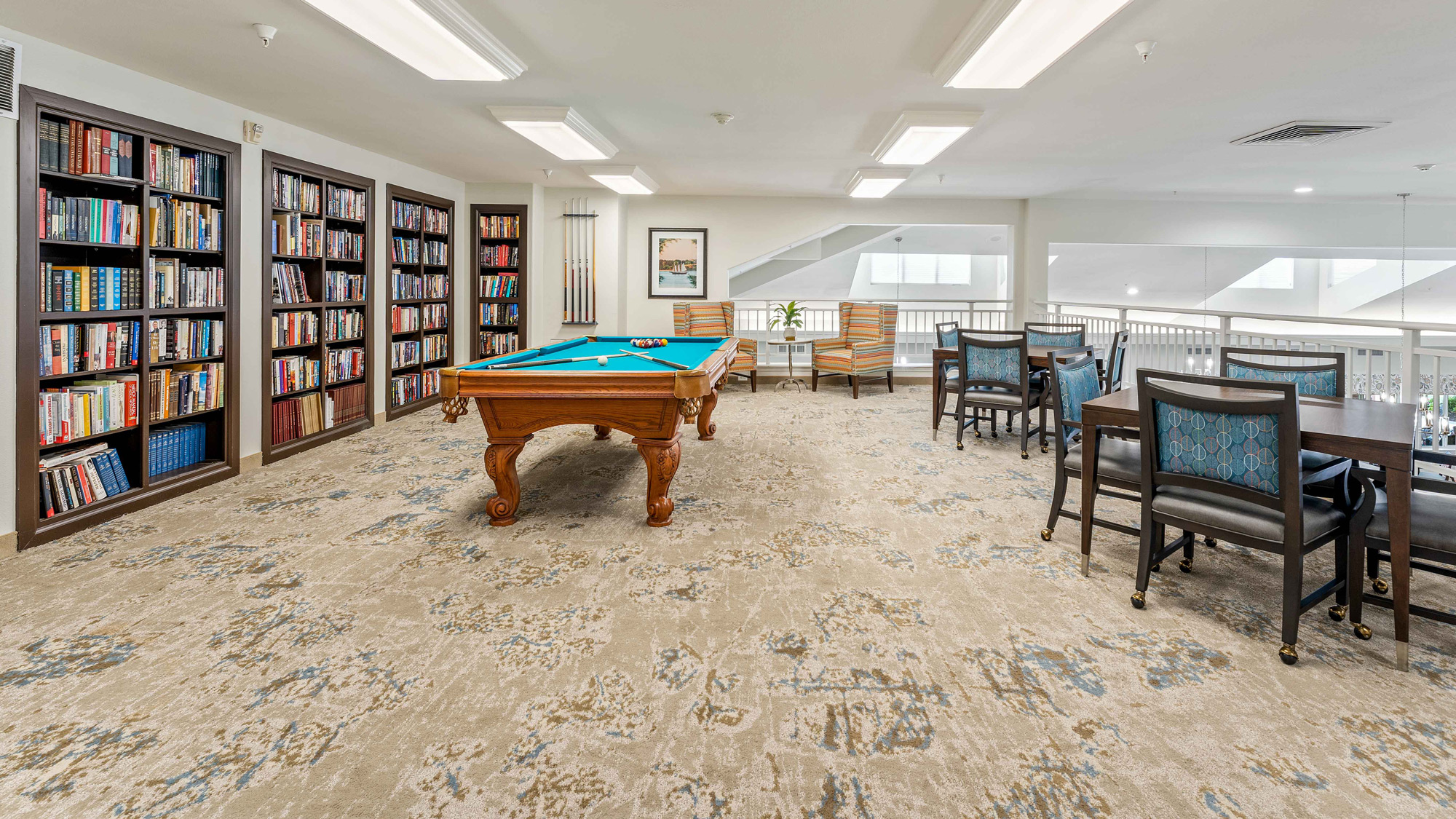 A spacious room with bookshelves along the wall, a pool table in the center, and a seating area with tables and chairs near a railing overlooking a lower floor. The room is well-lit with overhead lights.