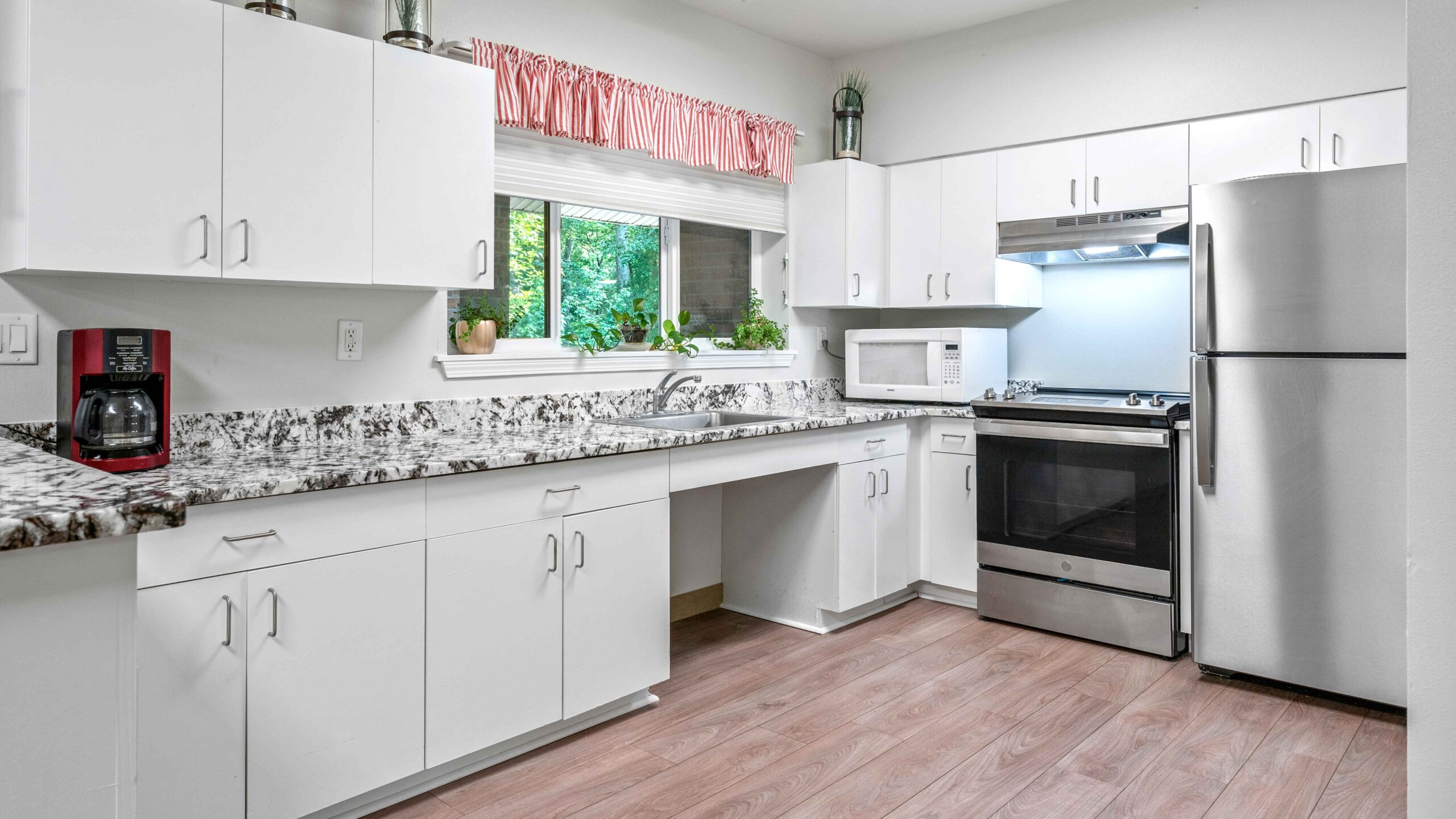 Modern kitchen with white cabinets, a stainless steel oven, refrigerator, microwave, a coffee maker, granite countertops, and a window with a pink valance above the sink. Light wood flooring completes the space.