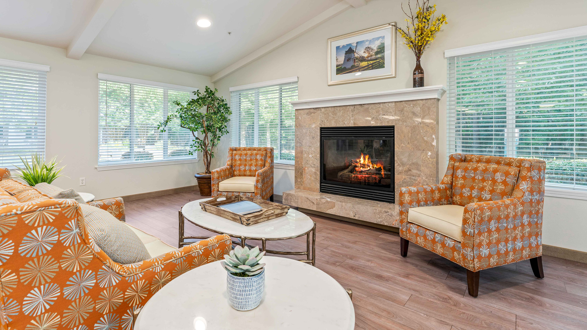 A cozy living room with orange patterned armchairs, a beige couch, round marble coffee tables, a potted plant, and a fireplace beneath a painting. Large windows bring in natural light and greenery from outside.