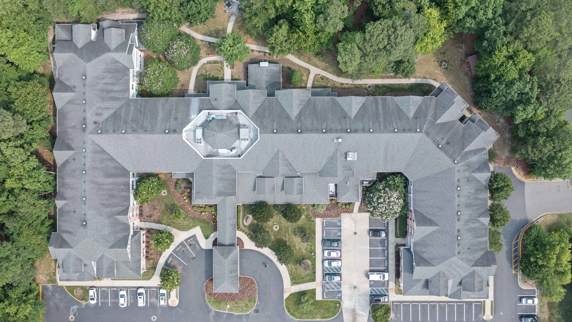 Aerial view of a large, gray-roofed building surrounded by trees, landscaped pathways, and parking lots with several parked cars. The building has a central octagonal section and multiple wings extending outward.