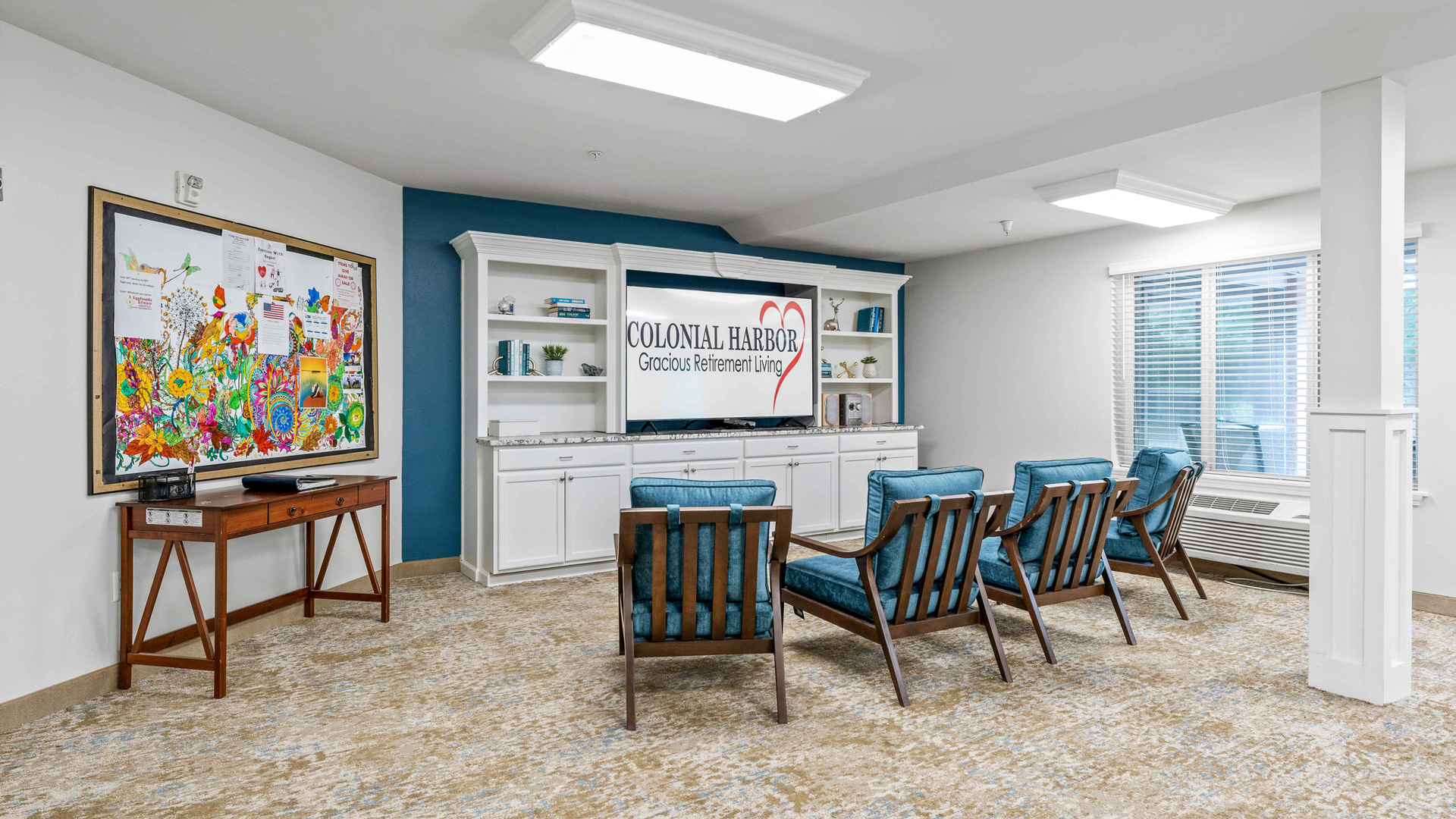 A bright lounge area with four blue cushioned chairs facing a TV, white built-in shelves, a window with blinds, and a colorful bulletin board on the wall with a wooden console table beneath it.