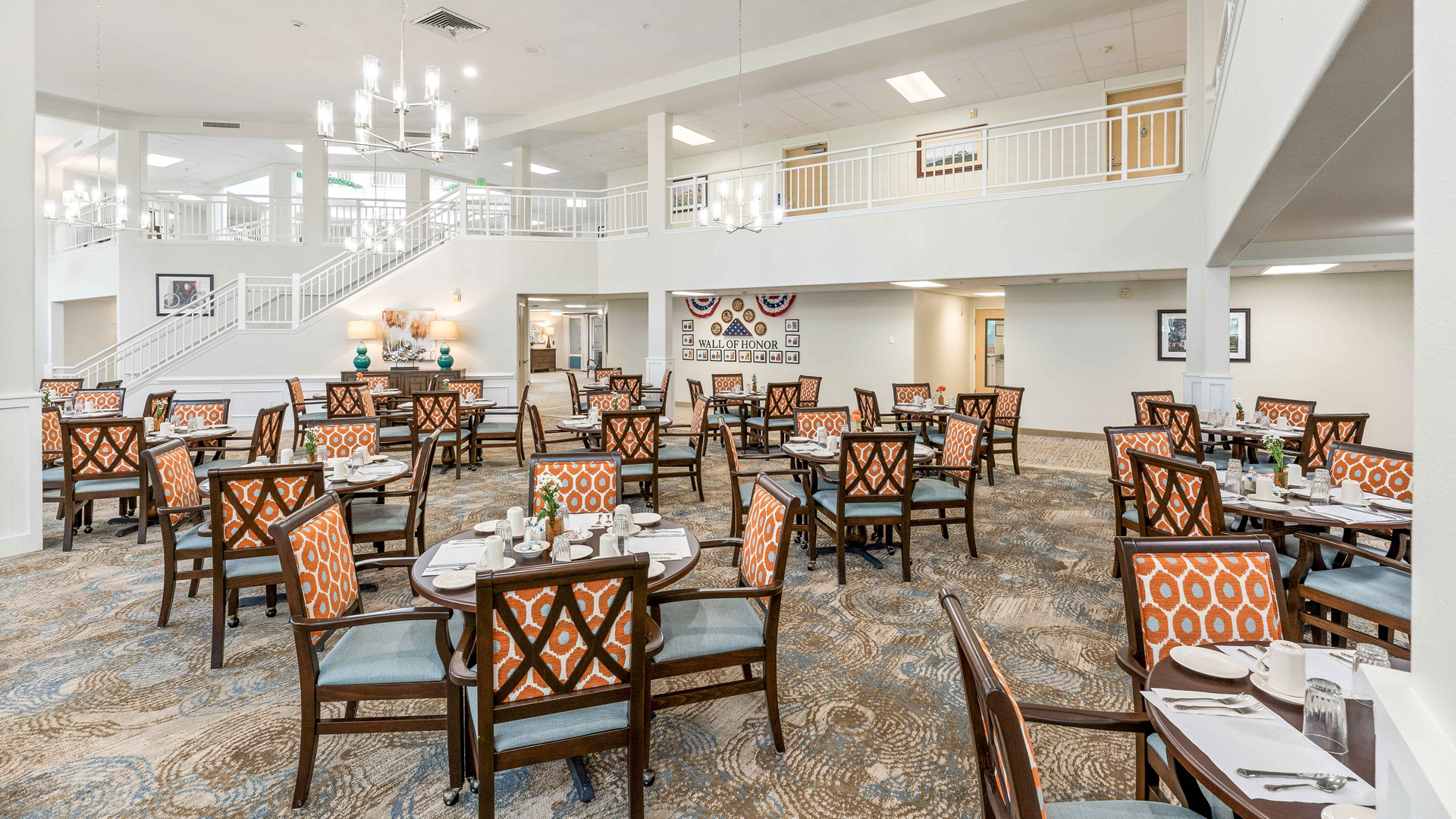 A spacious, well-lit dining room with neatly set round tables and cushioned chairs with orange patterned backs. There is a staircase, a mezzanine, and large windows contributing to an airy, inviting atmosphere.