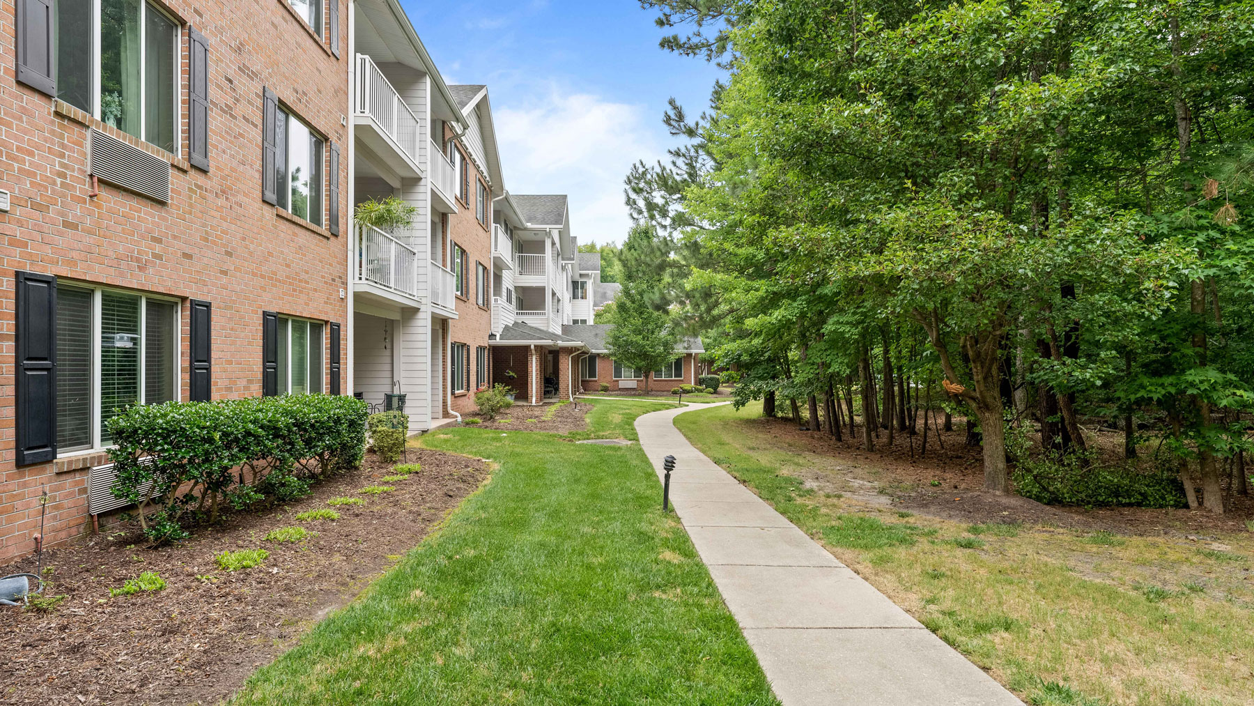 A sidewalk curves between a brick apartment building with balconies on the left and a wooded area with green trees on the right, under a clear blue sky.